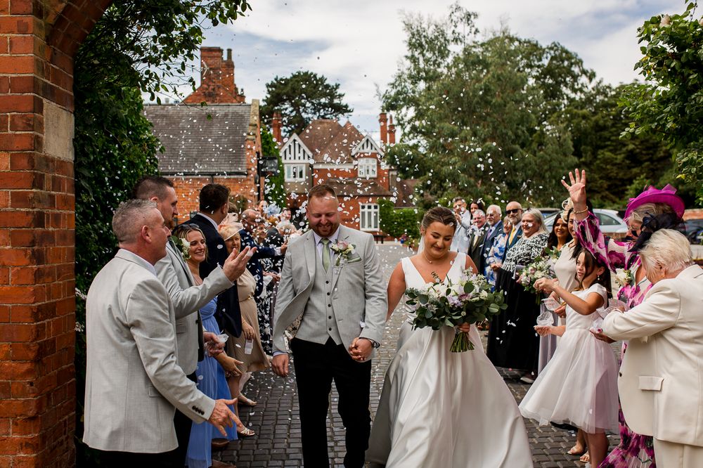 Wedding guests throw confetti at a newly married couple during an outdoor celebration at a brick venue.