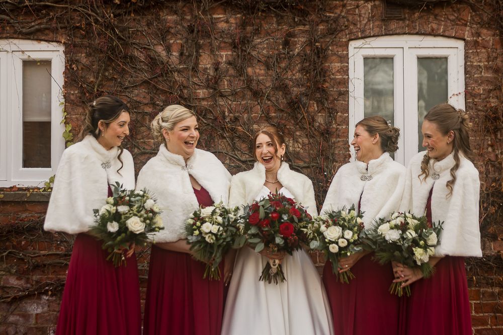 Bridesmaids in burgundy dresses and white sweaters pose together against a rustic brick wall with winter bouquets.