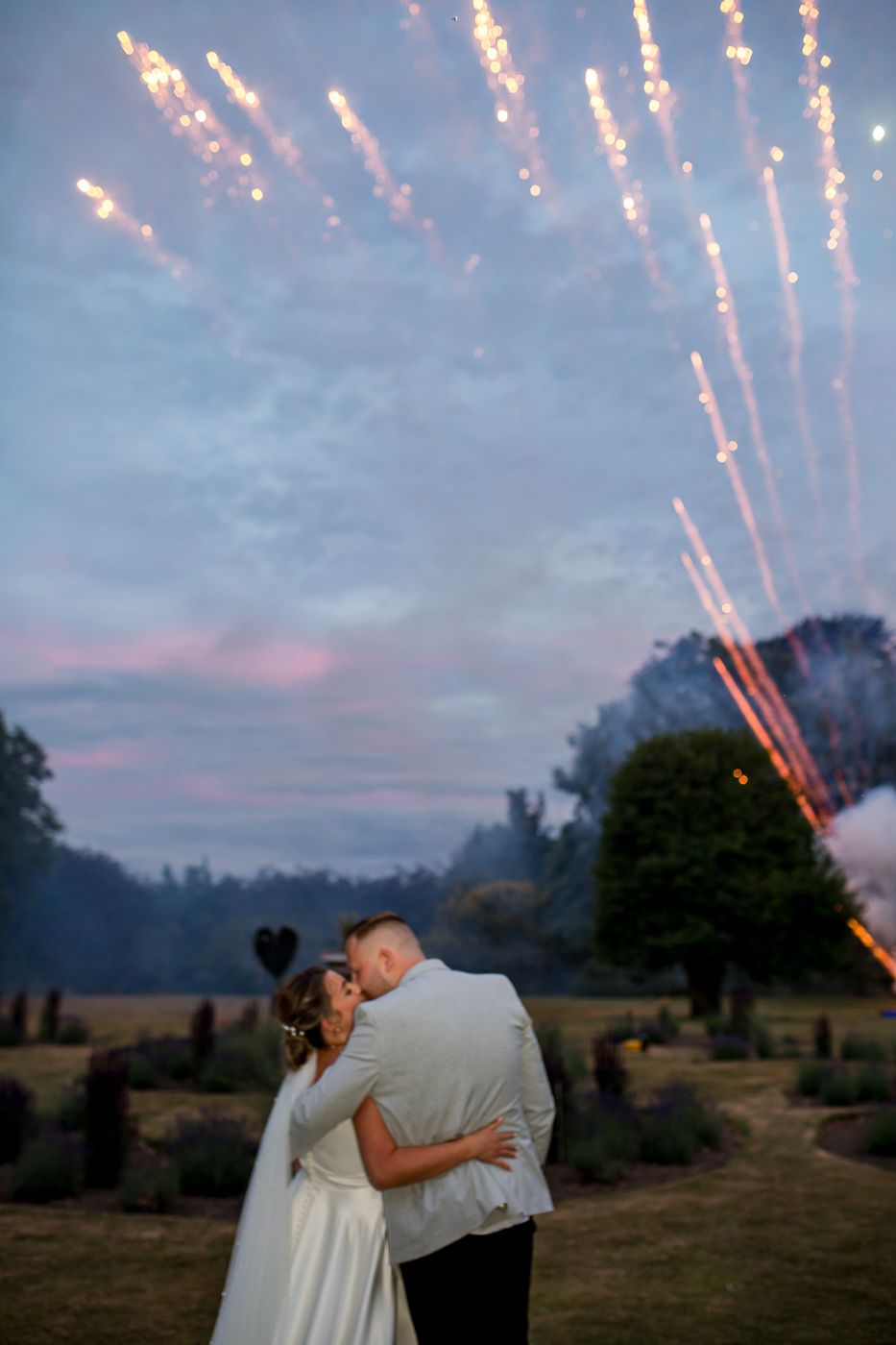 A couple embraces under a dramatic sky filled with fireworks creating a romantic silhouette against the twilight landscape.