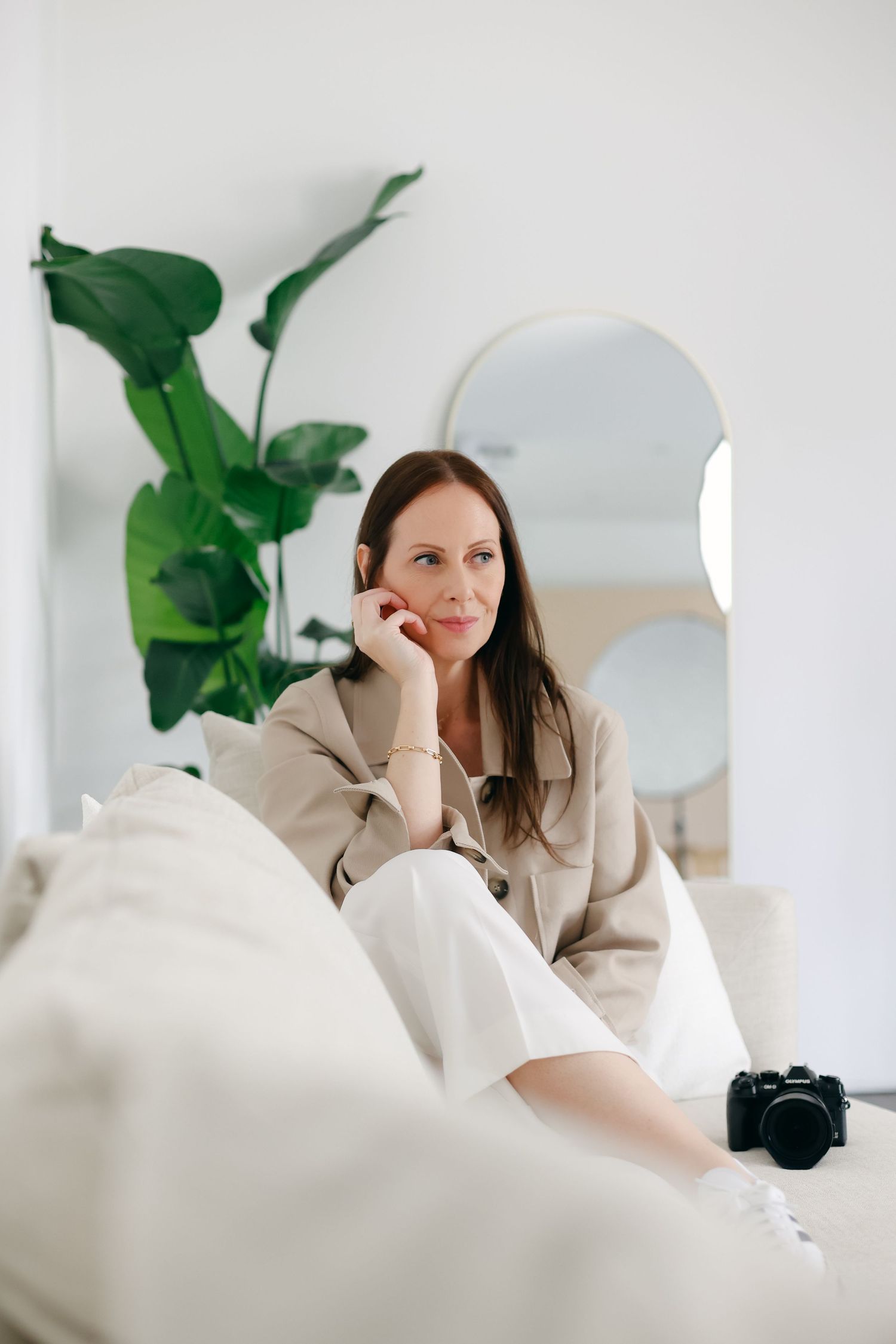 A relaxed person in a beige sweater sits on a white couch near a large houseplant and digital camera.