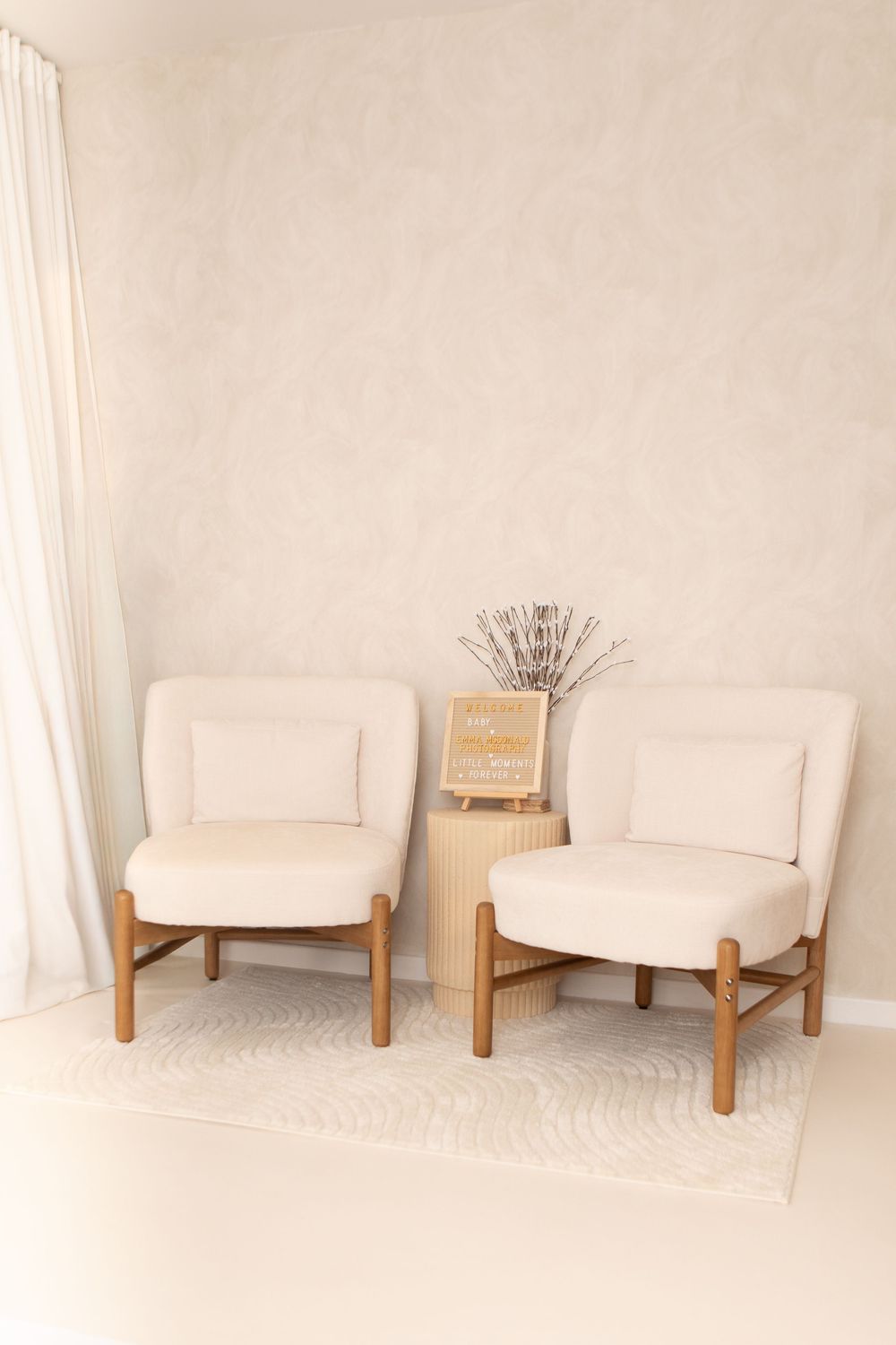 Two cream-colored accent chairs with wooden frames and a small table with dried flowers between them on a light carpet.