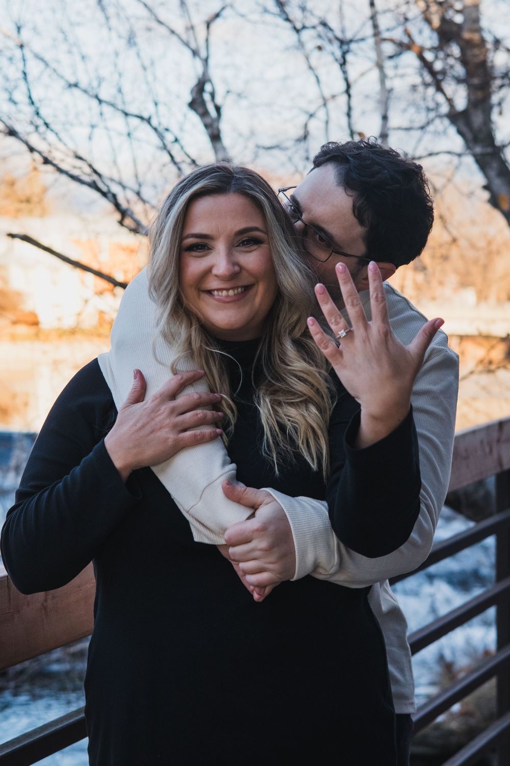 A couple shares a playful embrace on a winter day with bare trees in the background.