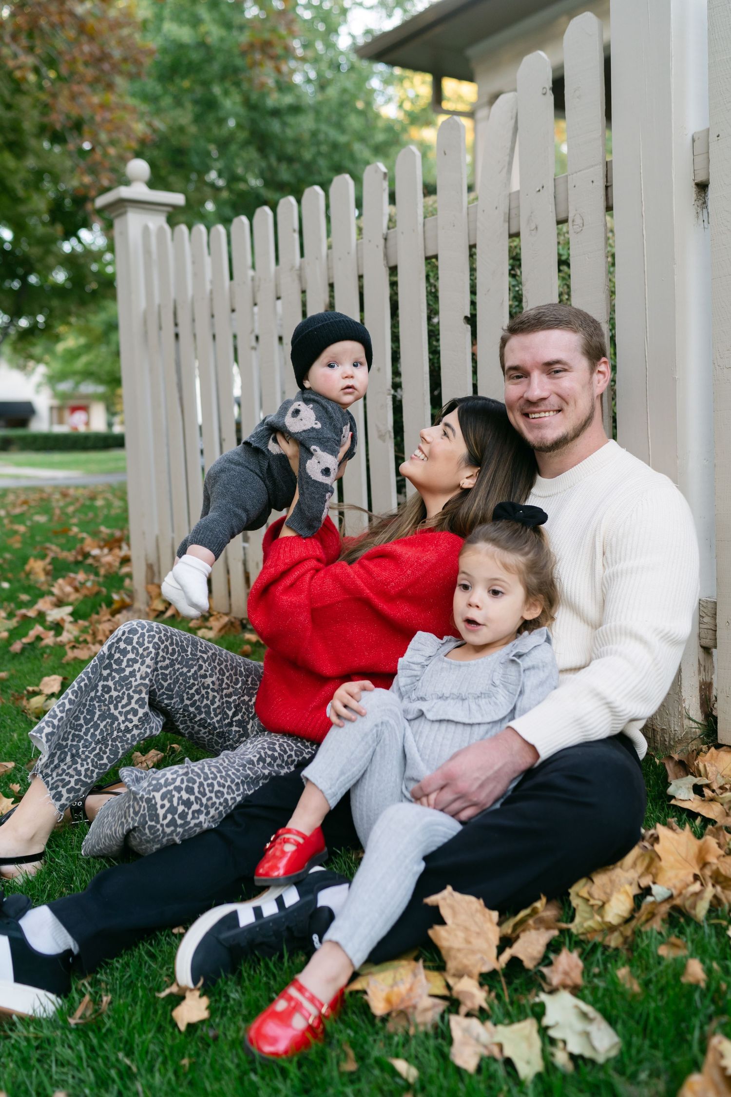Family relaxing on grass with fallen leaves by white picket fence.