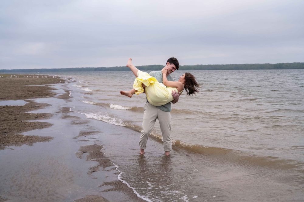 Couple dancing joyfully on a sandy beach shore, man lifting woman in yellow dress with ocean waves in background.