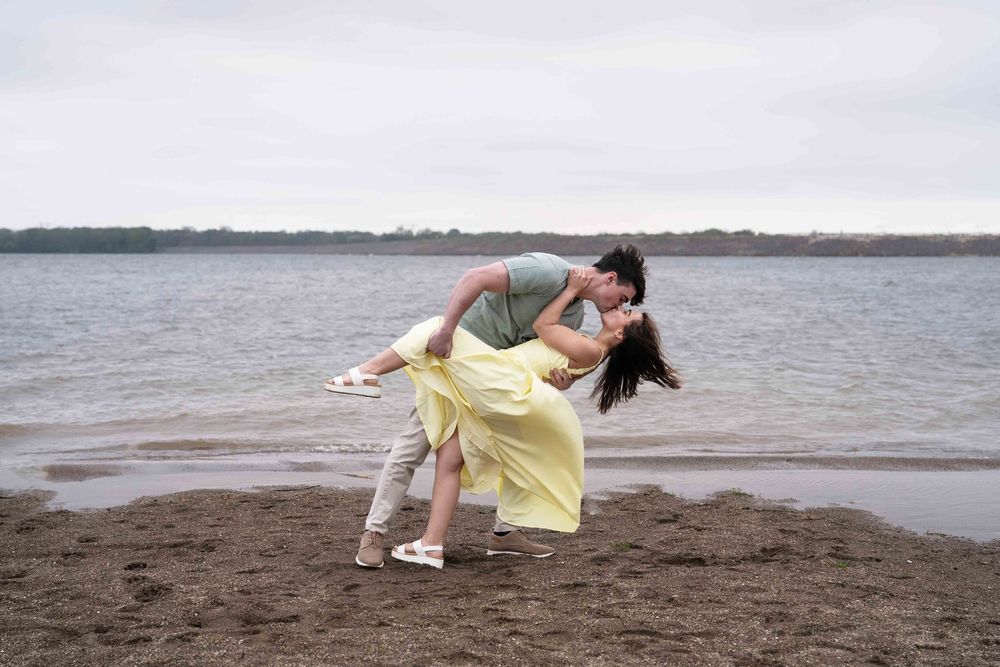 Romantic couple sharing a dip kiss on a sandy beach shore with calm lake waters in the background.