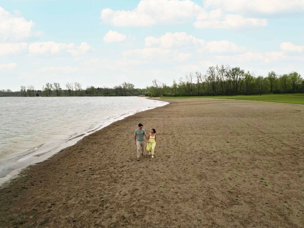 Couple holding hands walking along a sandy beach shoreline with trees and cloudy sky in the background.
