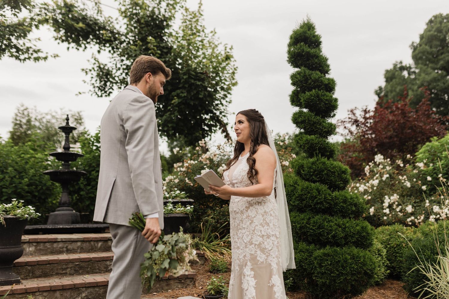 A romantic wedding ceremony takes place in a garden setting with manicured topiary and stone steps.