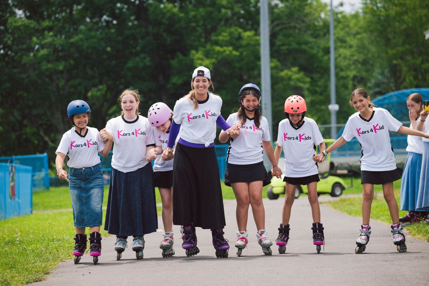 Group of girls roller skating together outdoors with instructor, wearing matching white t-shirts and helmets on a sunny day.
