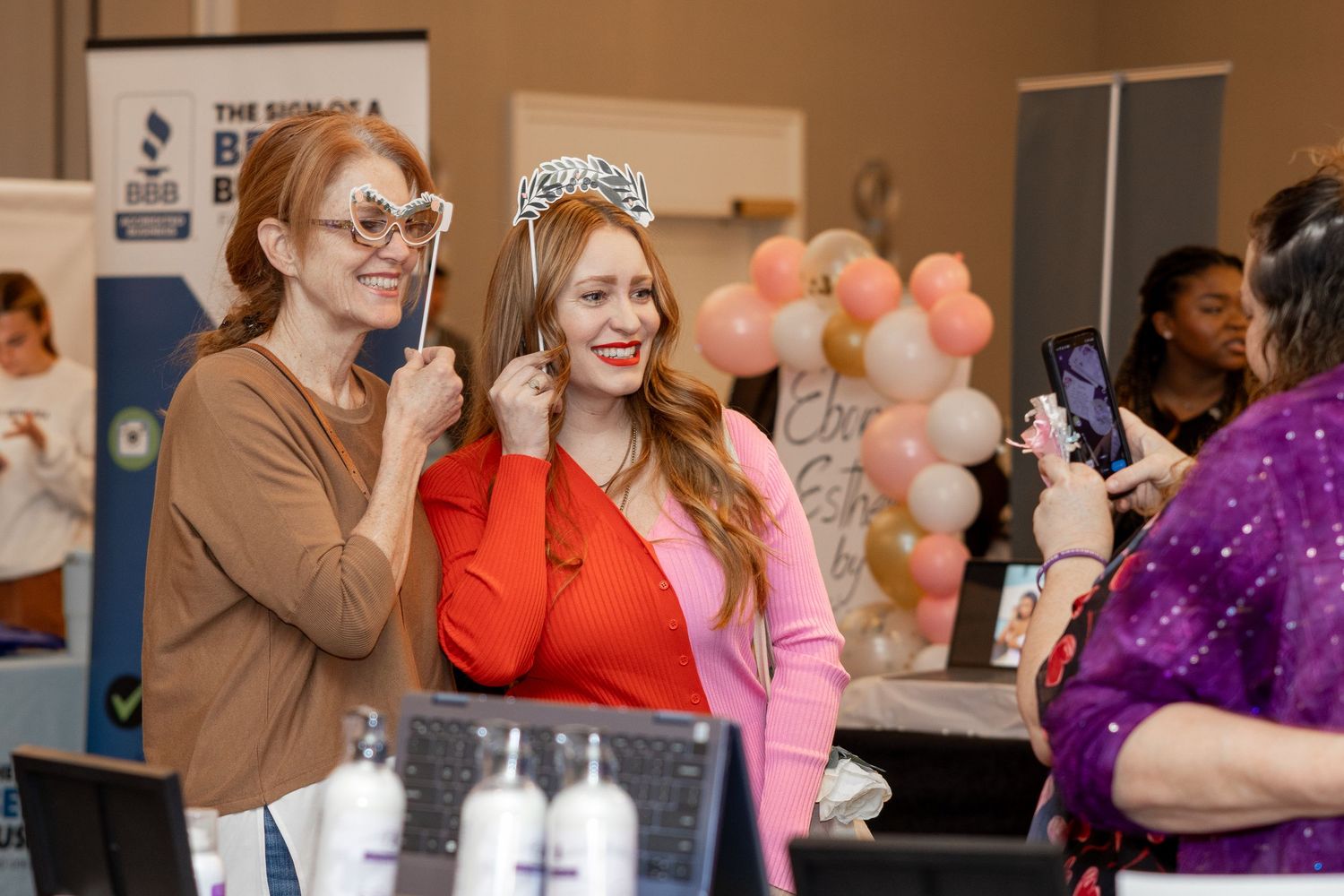 Two attendees share a joyful moment at an event with decorative balloons in the background.