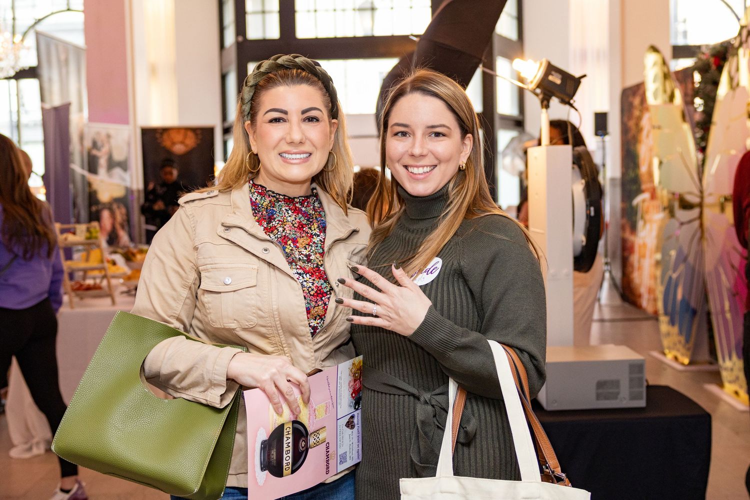 Two shoppers with handbags smile while browsing at an indoor retail event.