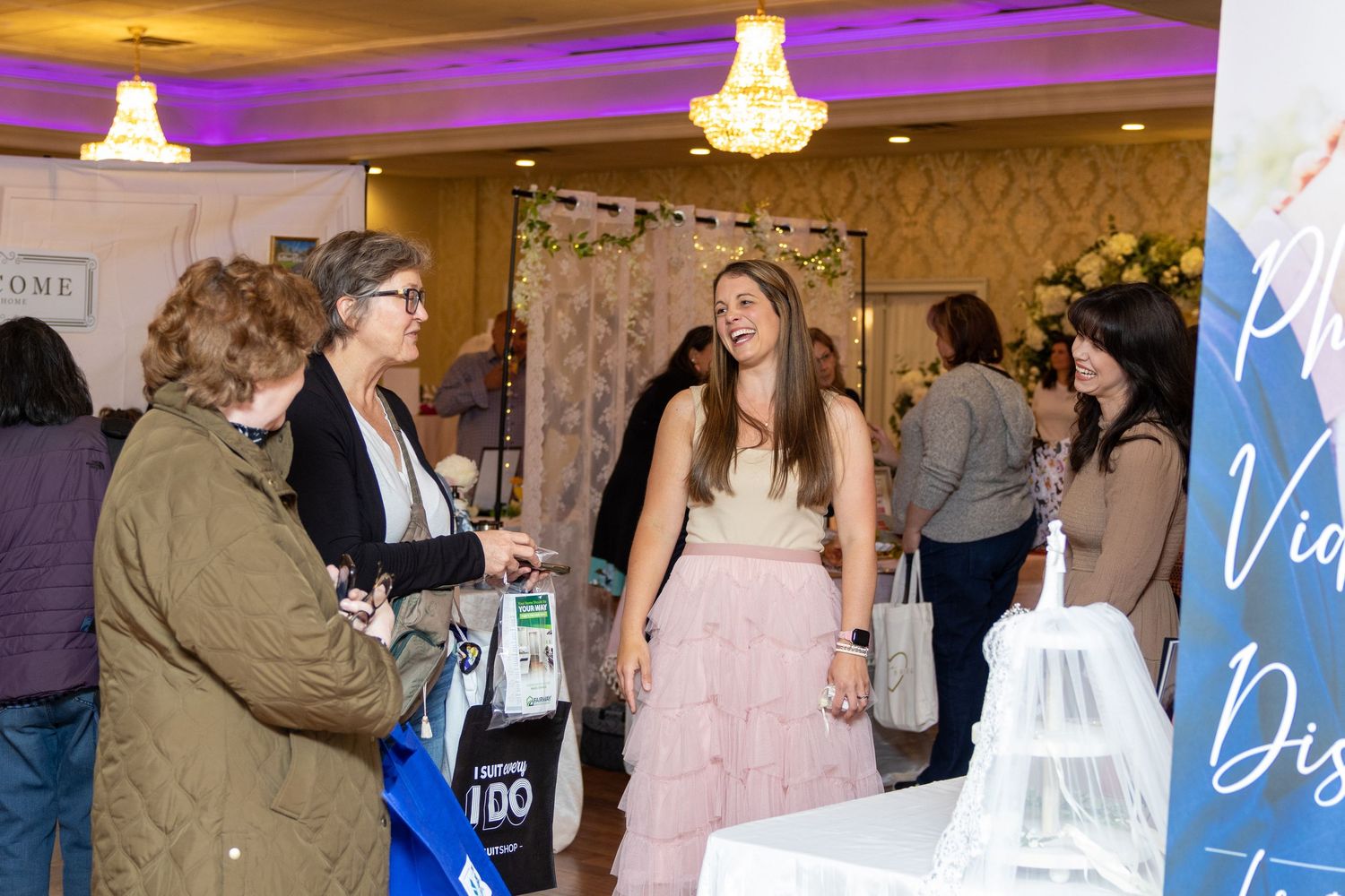 Attendees interact at a bridal or wedding expo with purple mood lighting and chandeliers.