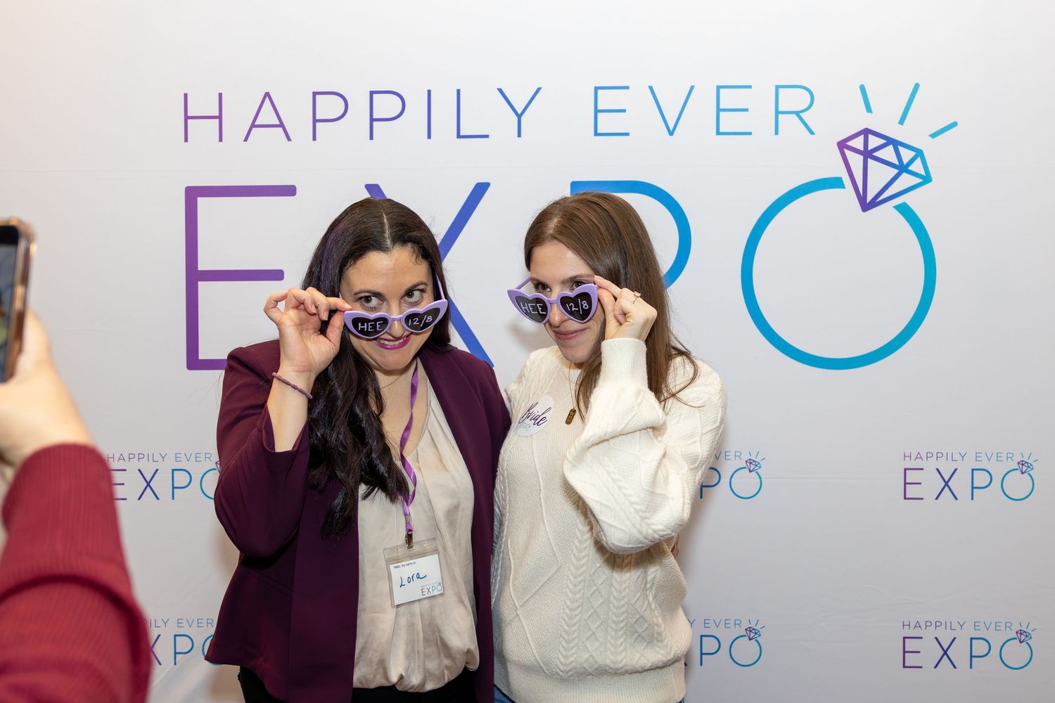 Two attendees pose playfully with sunglasses at a wedding expo photo wall.