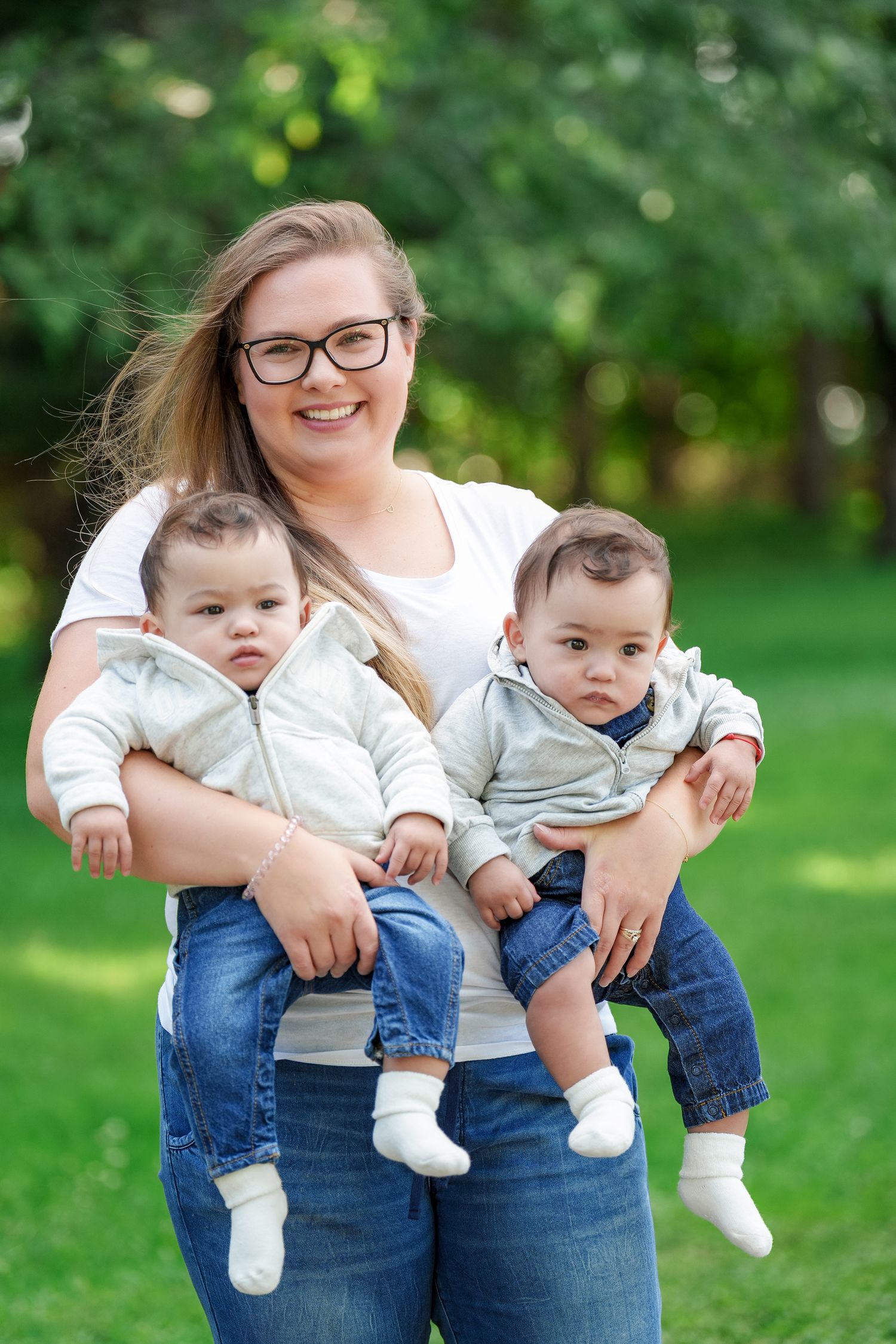 Three candid outdoor photos of a woman in glasses sitting with a baby in jeans and white shirt against a green background.
