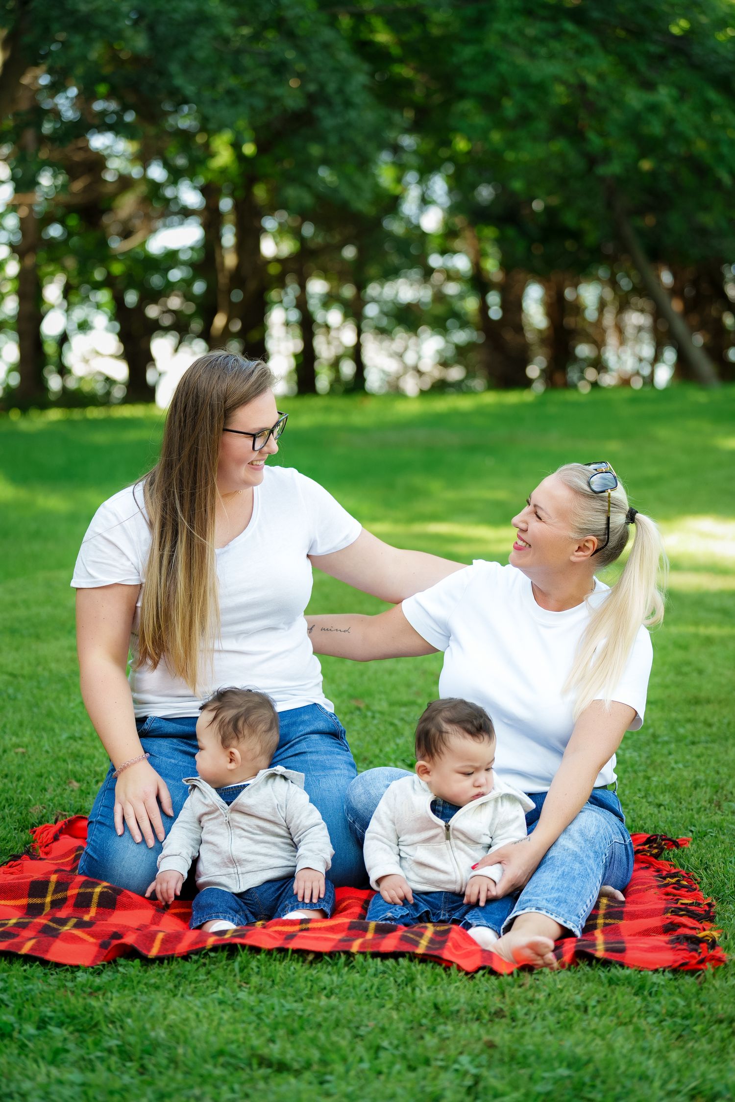 Family relaxing together on a red picnic blanket in a sunny green park setting.