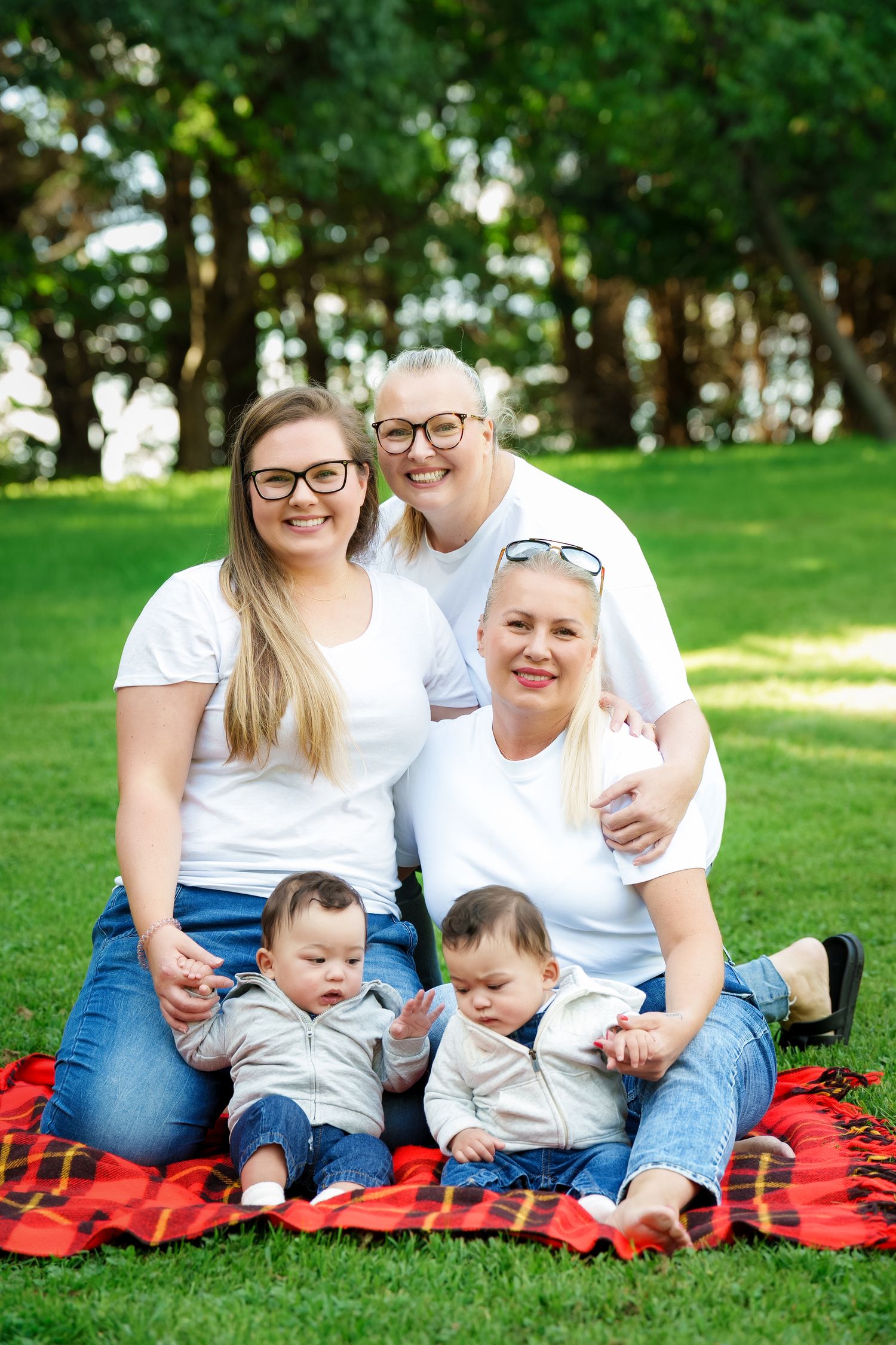 Casual outdoor family portrait with everyone wearing white shirts and sitting together on red blanket in park.