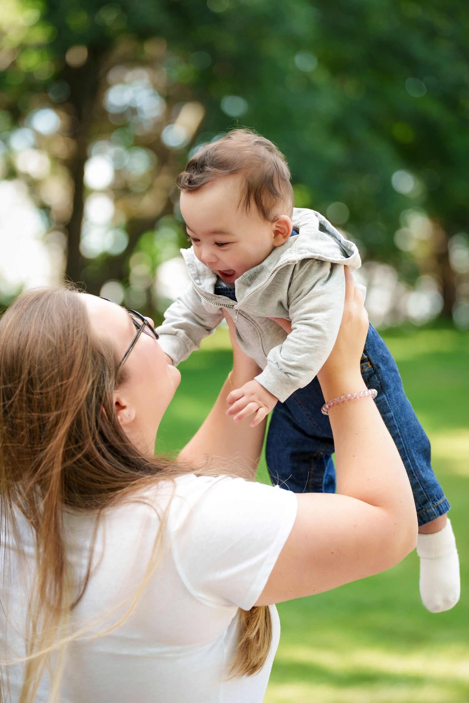 A joyful moment captured outdoors as a baby is lifted high in the air on a sunny day in a green park setting.