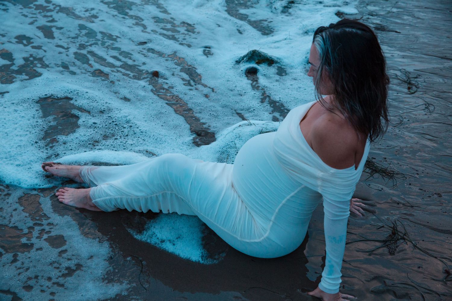 Figure in a light blue dress poses on wet sand during blue hour at the beach.