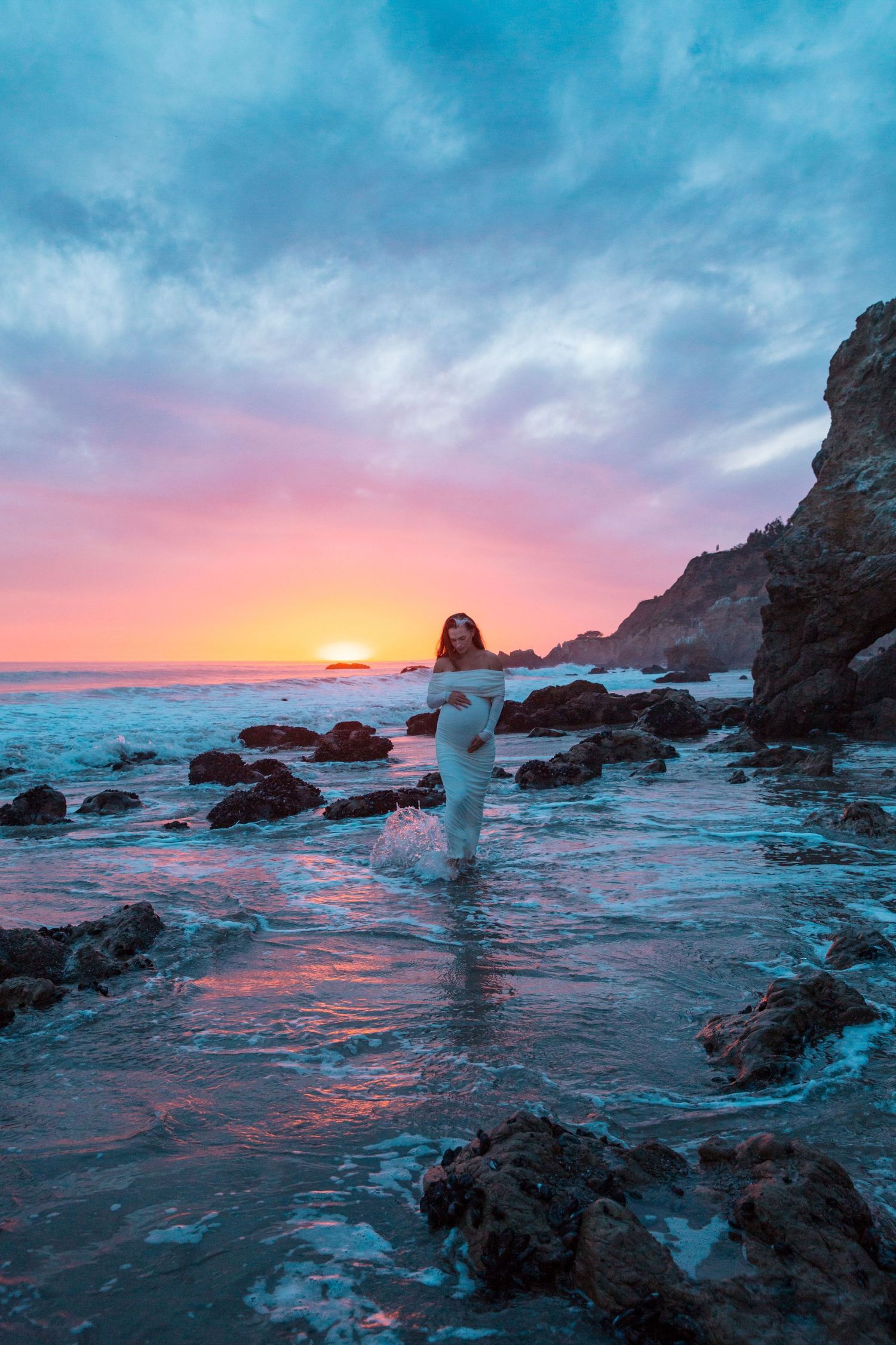 Silhouetted figure stands in shallow ocean water against dramatic pink and orange sunset with rocky coastline.