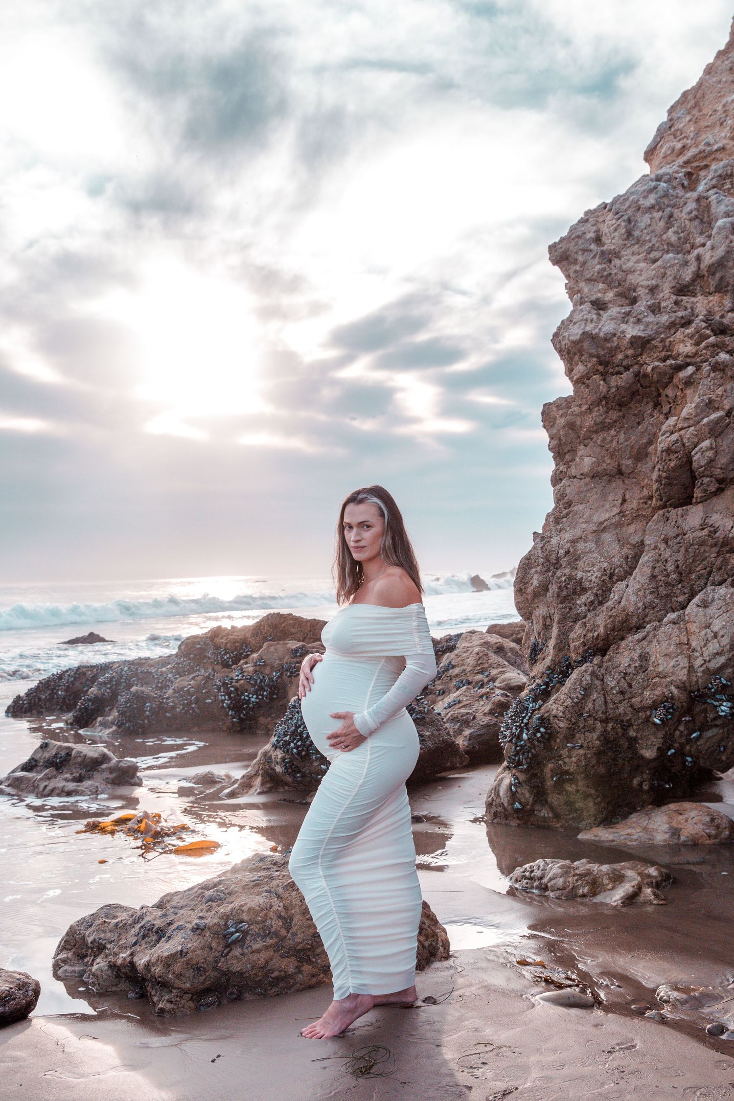 A pregnant woman in a white dress poses by coastal rocks at sunset on a misty beach.