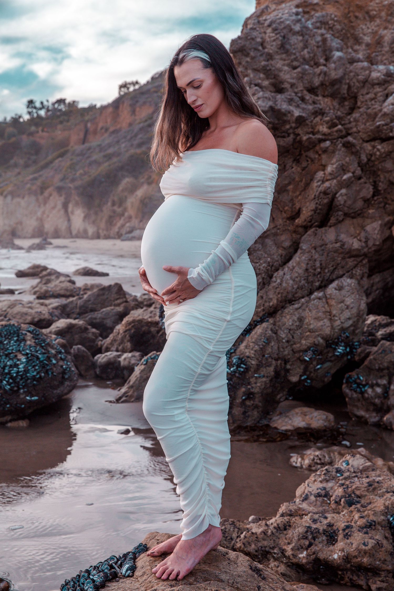 A maternity photoshoot on a rocky beach shore features an elegant white dress against dramatic coastal cliffs at sunset.