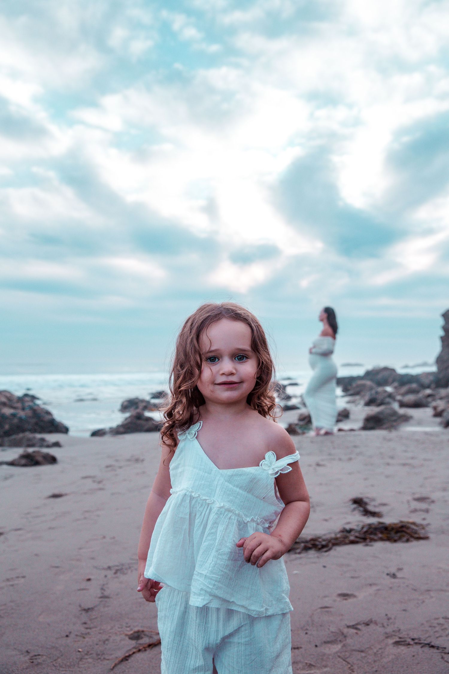 Child in flowing mint dress stands on rocky beach at sunset with cloudy blue sky and ocean waves in background.