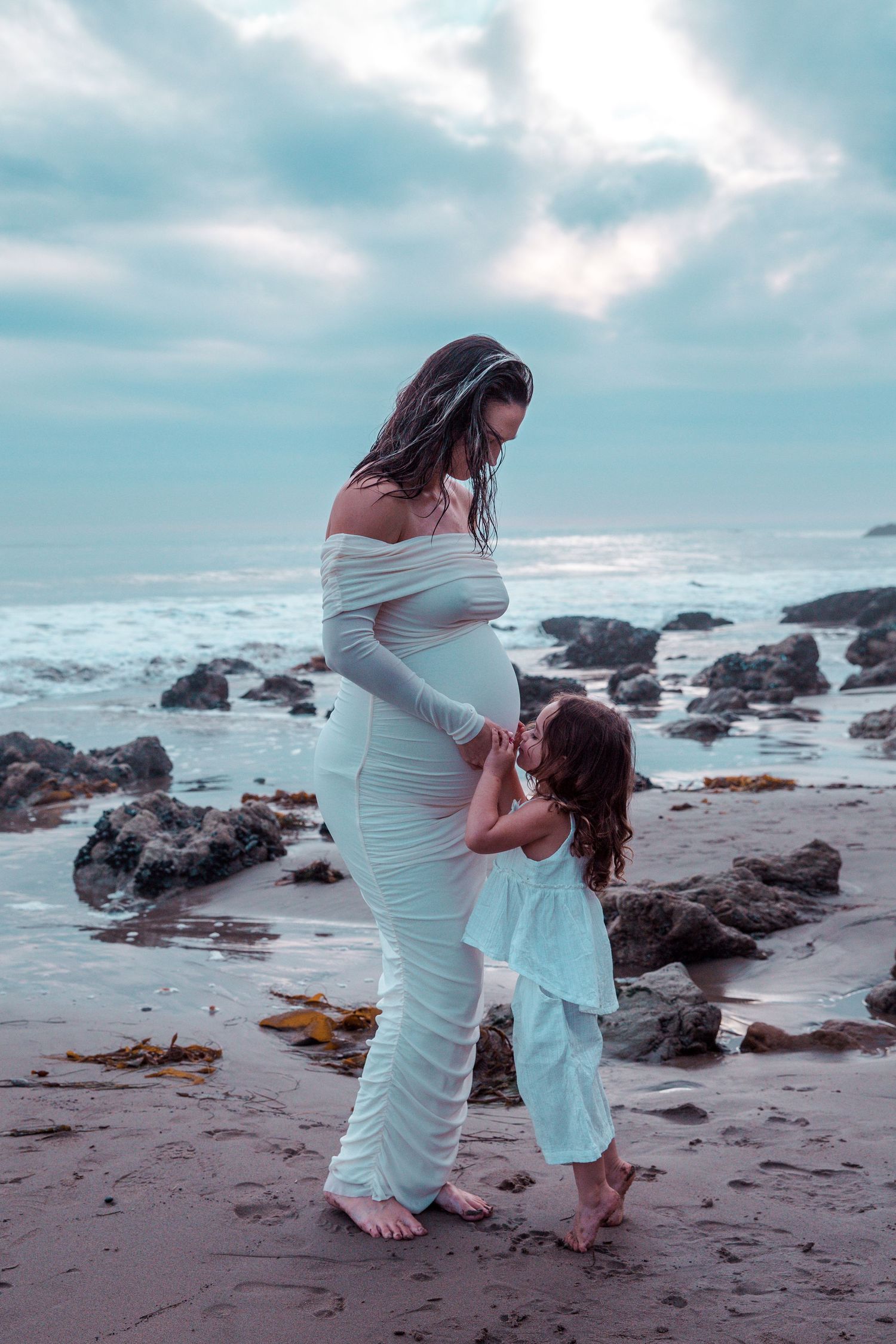 Touching seaside maternity photo with mother and daughter interacting on a rocky beach at dusk.