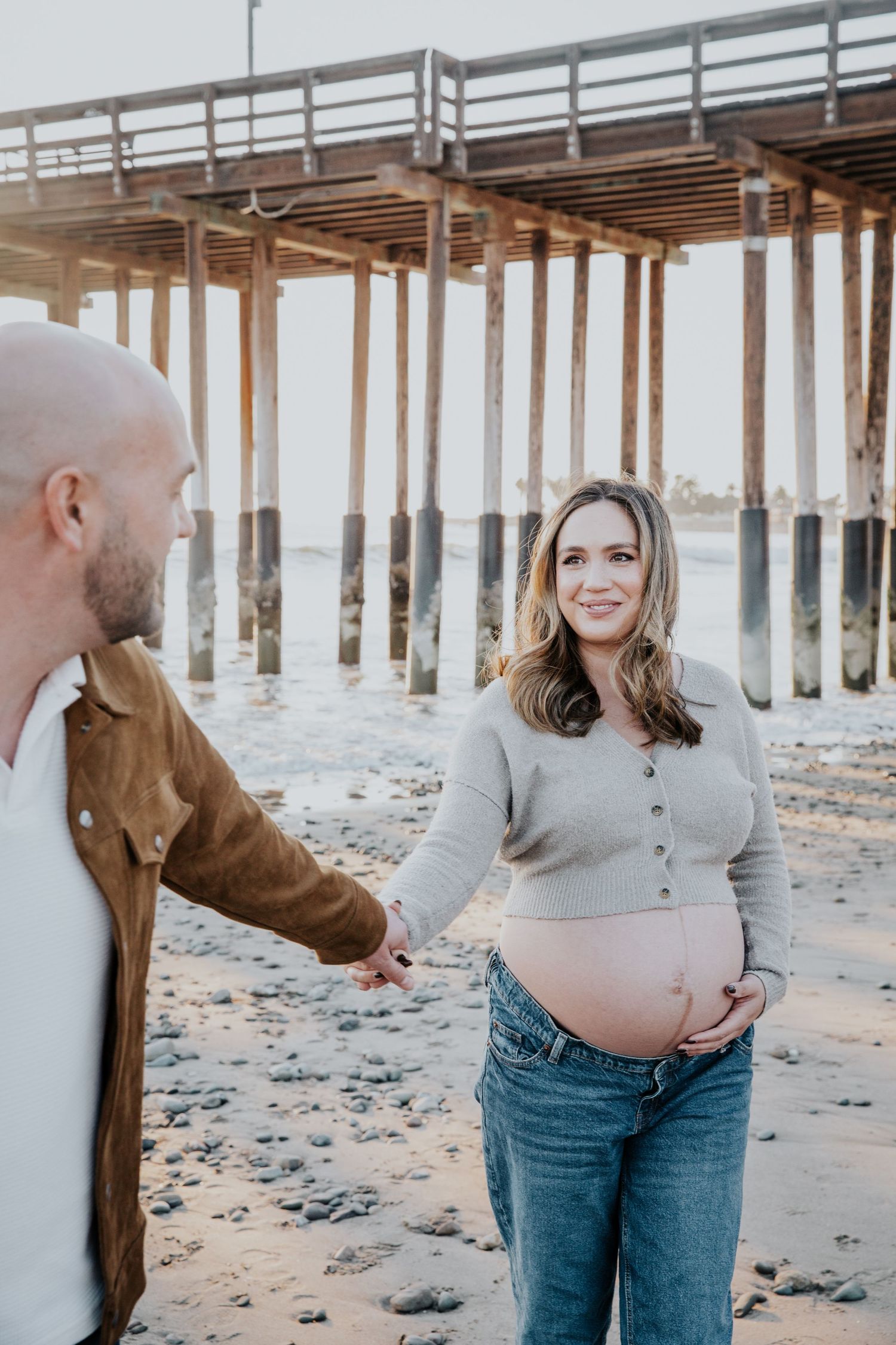 Expectant couple sharing a tender moment on the beach near a wooden pier.