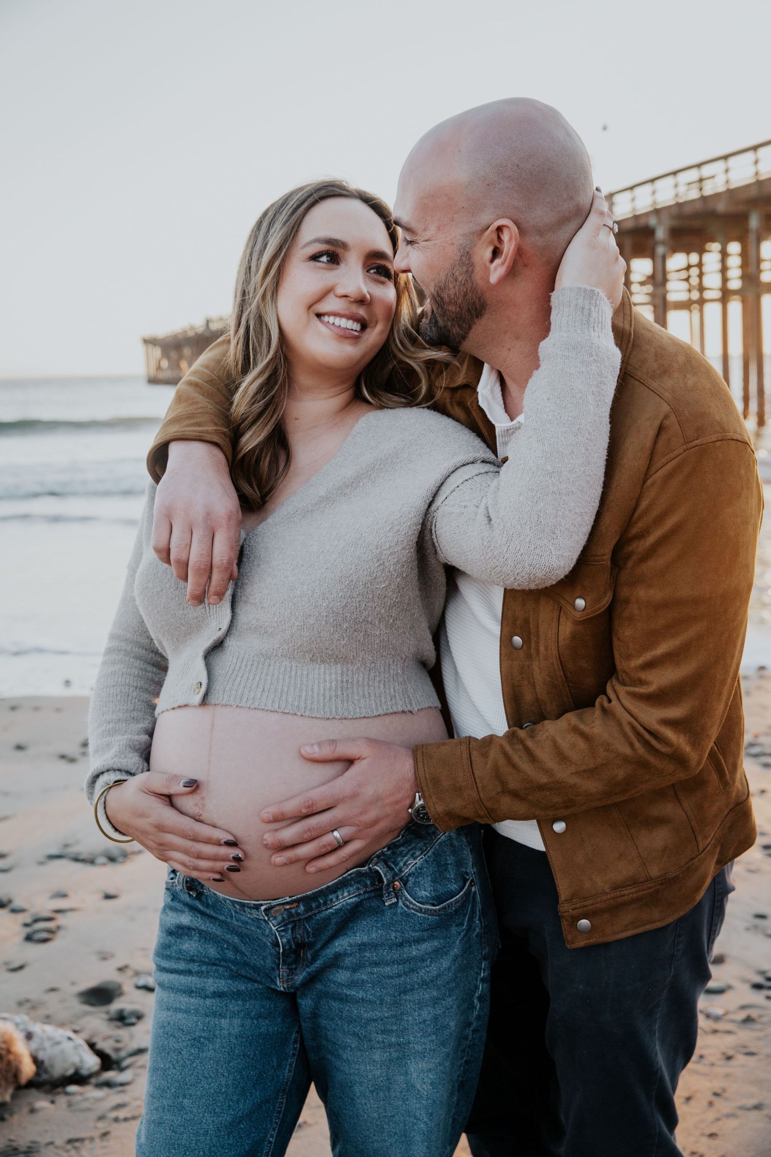 Intimate maternity photo of couple embracing on beach with wooden pier in background.