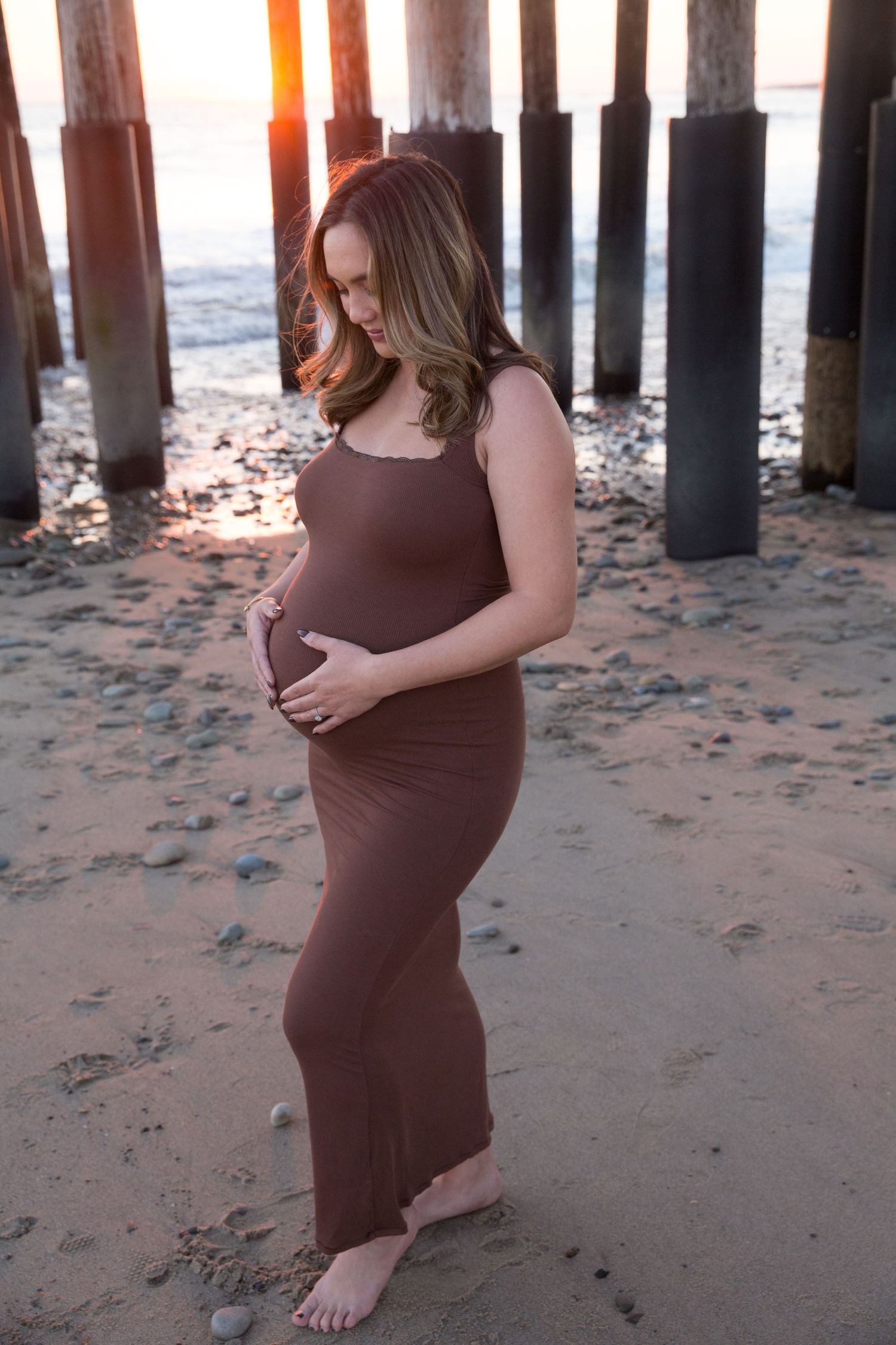 Silhouette of pregnant figure in brown dress standing beneath pier at sunset.