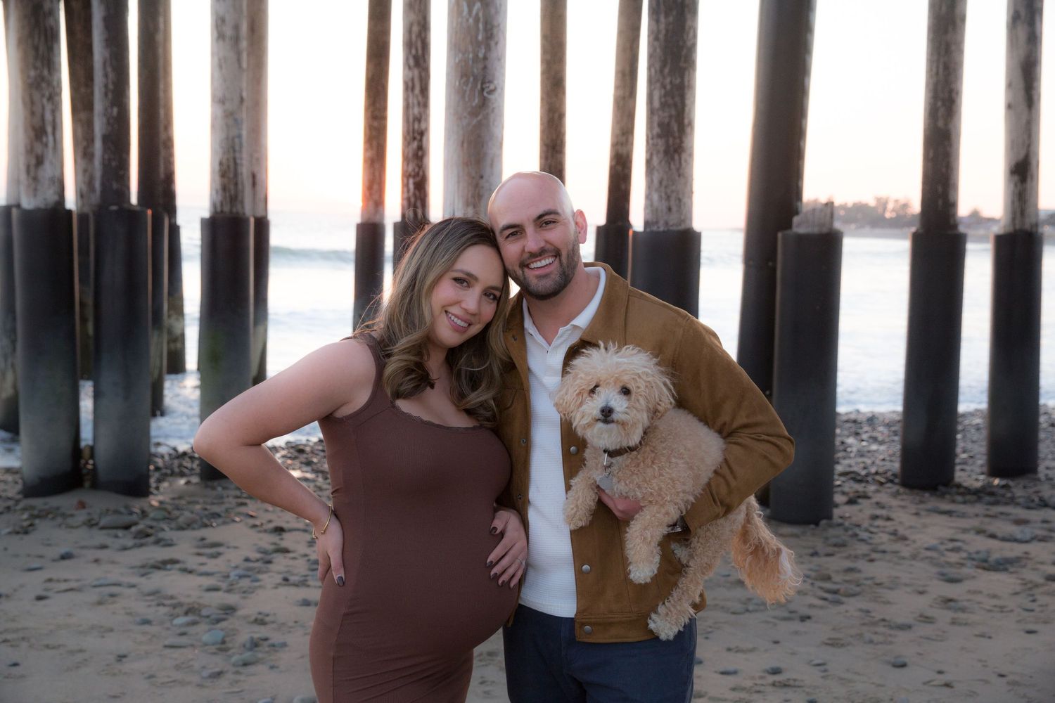 Maternity beach photo with couple holding small white dog under wooden pier.