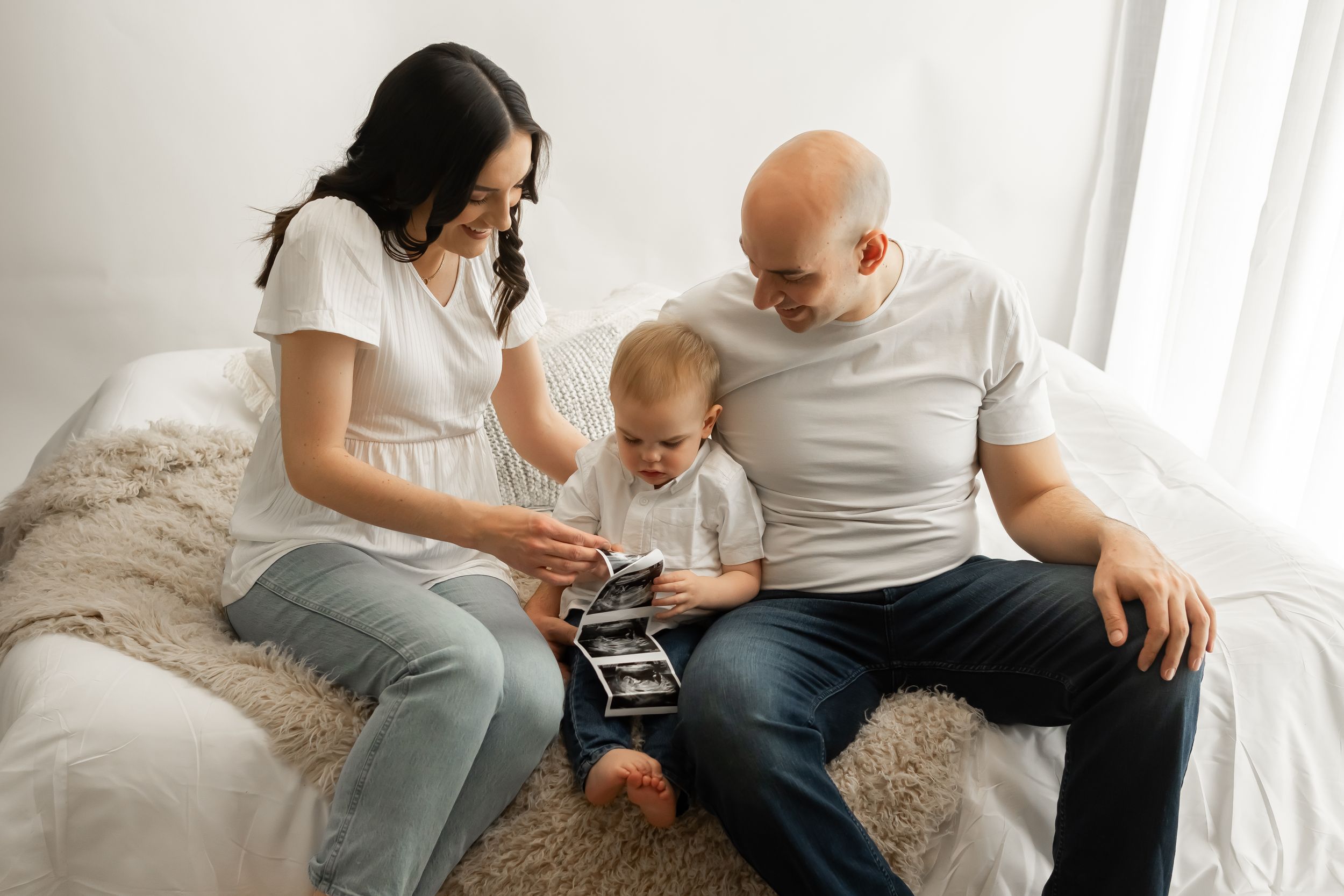 Parents and small child looking at ultrasound photos while sitting on bed.