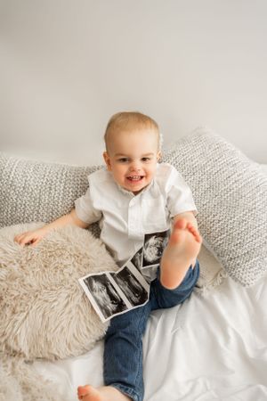 Toddler smiling while holding ultrasound photos on grey couch.