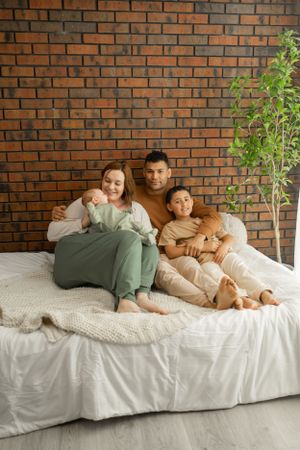 Family lounging together on white bedding against a brick wall with natural light and greenery.