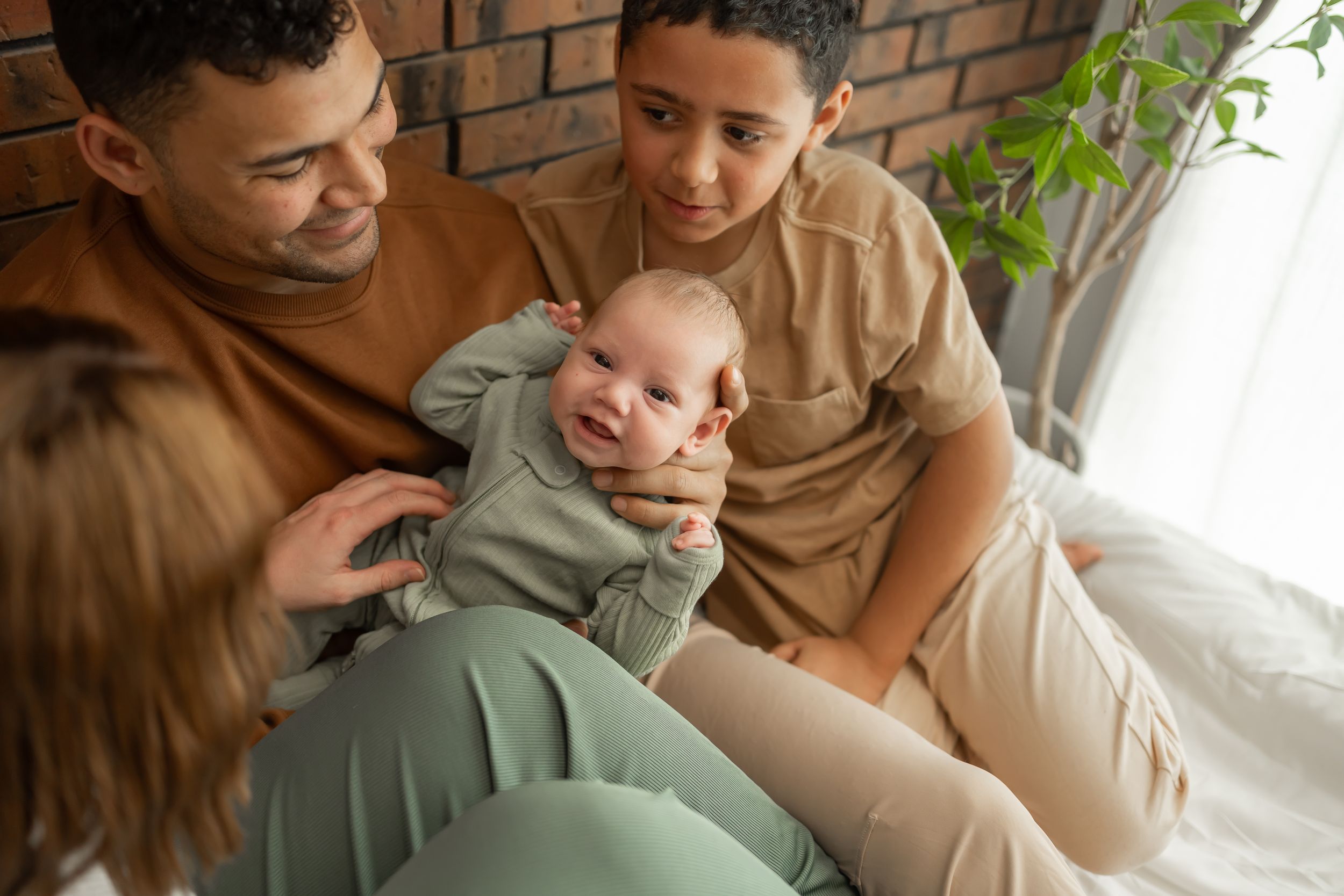 Tender family moments captured on white bedding against rustic brick background.
