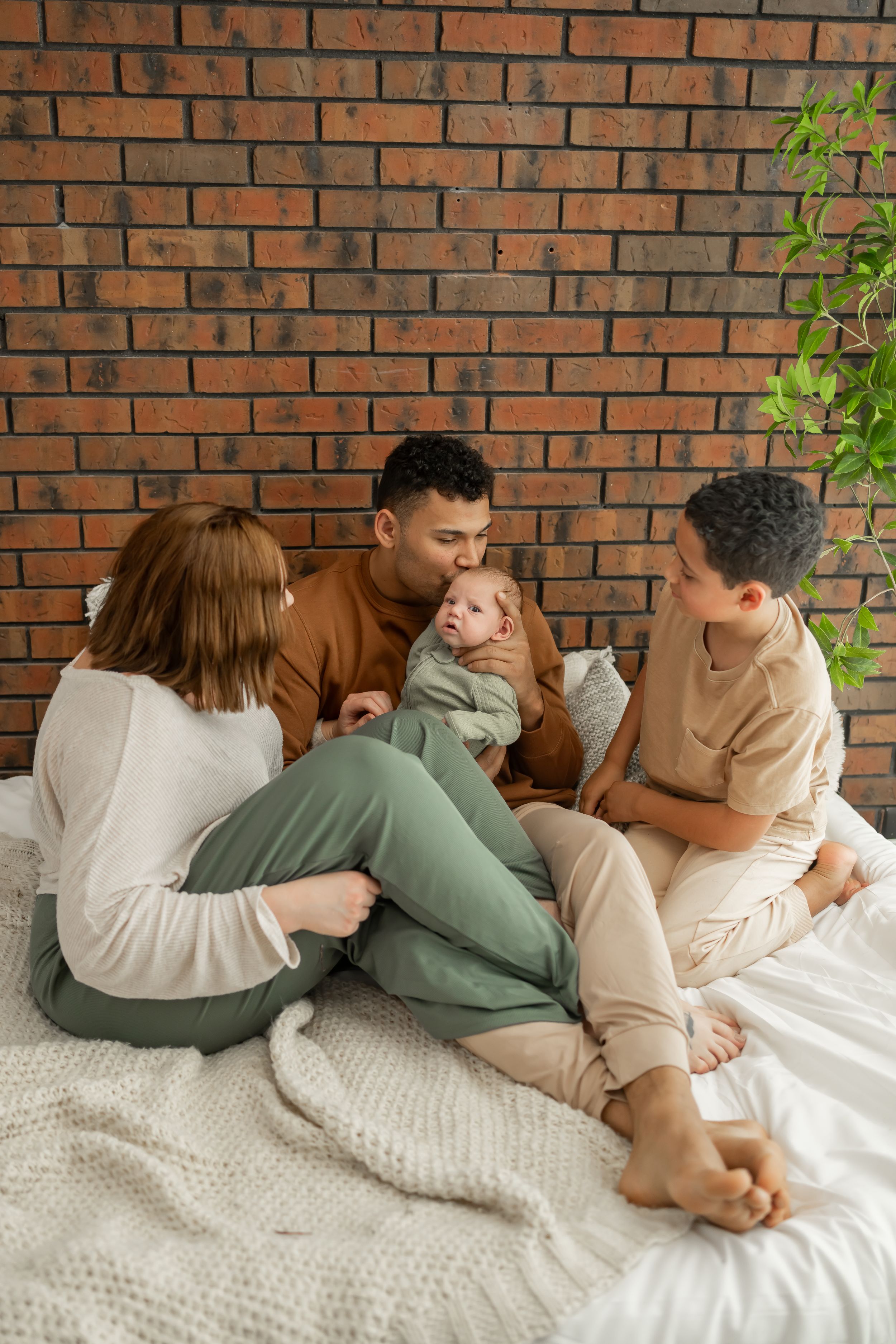 Family relaxing together on white knit blanket with exposed brick wall and window light.