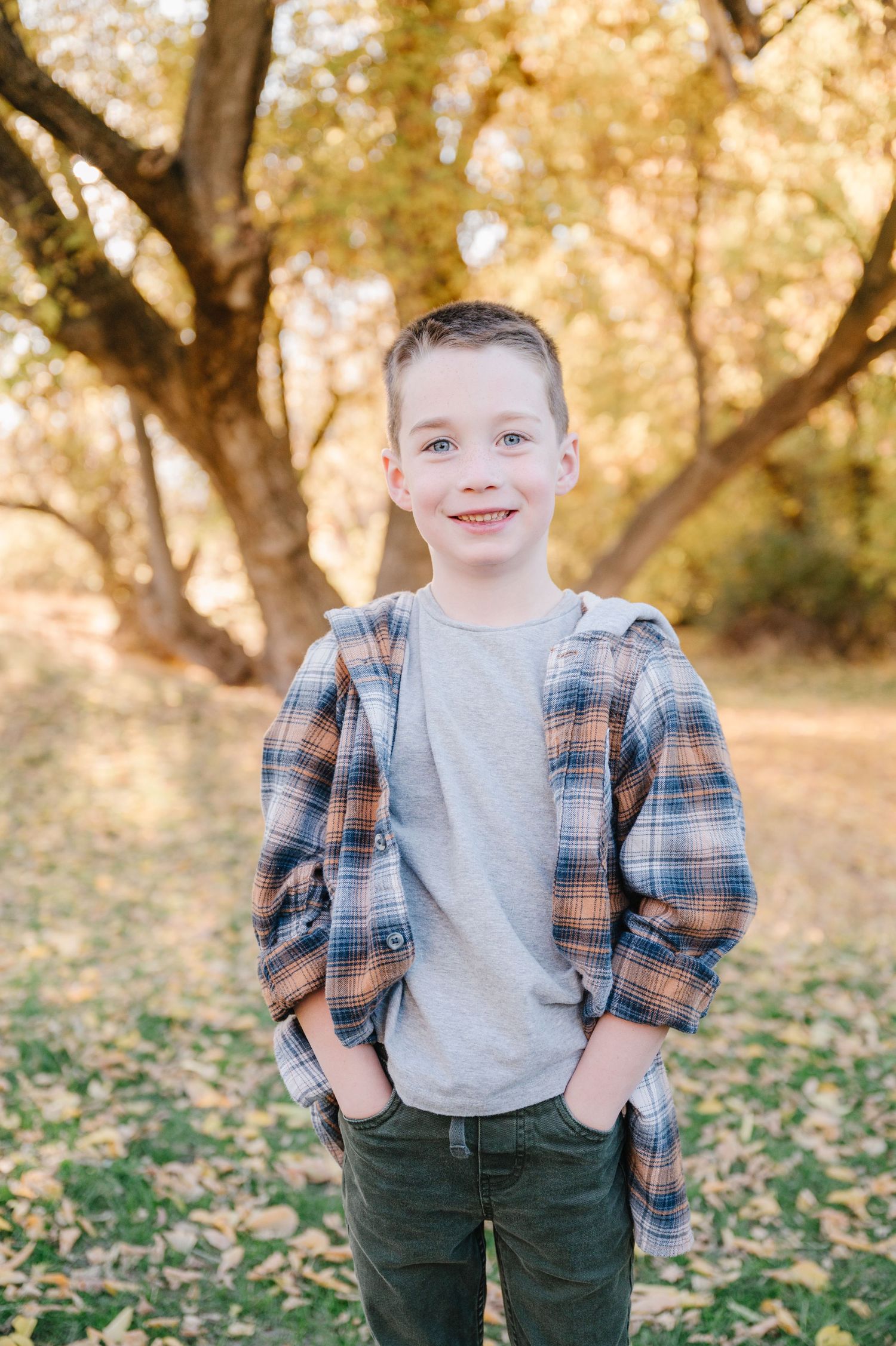 A young child wearing a plaid shirt and gray t-shirt poses outdoors among autumn trees and fallen leaves.
