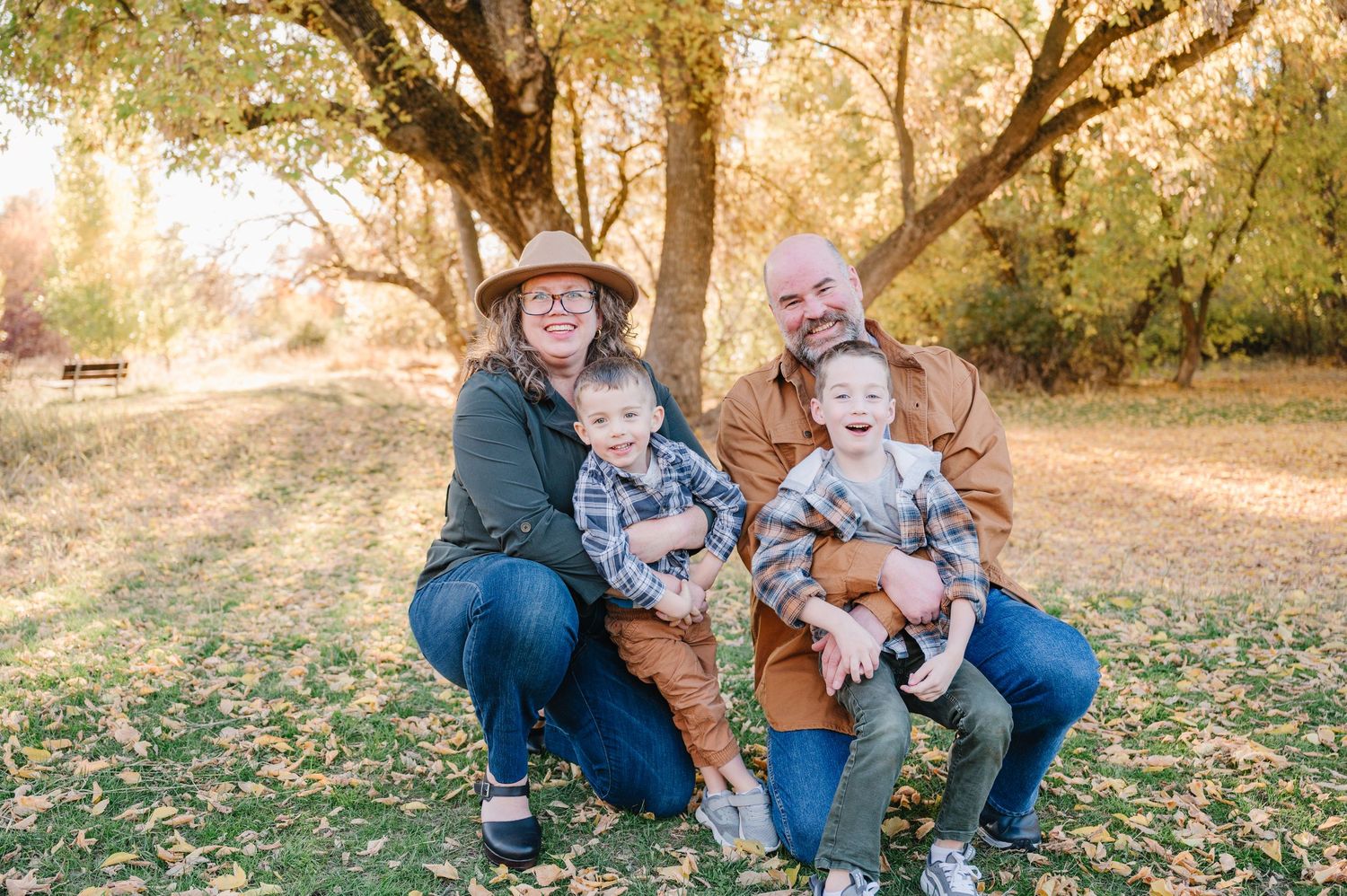 Family wearing coordinating blue and brown outfits sits together on fallen autumn leaves in a sunlit outdoor setting.
