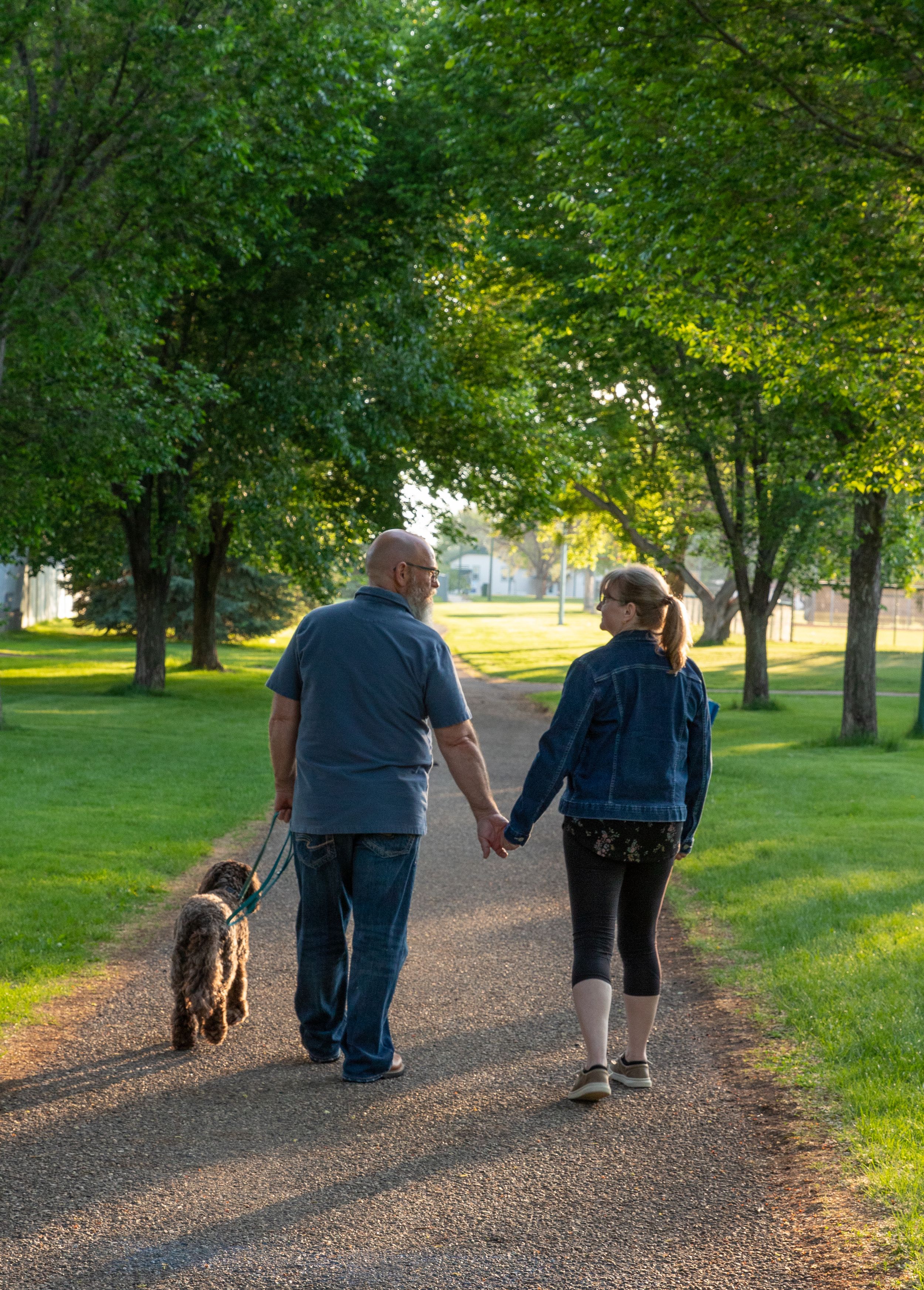 a couple in casual attire with a brown dog walking down a green tree lined path. Family lifestyle session in Alberta by Creation Photography