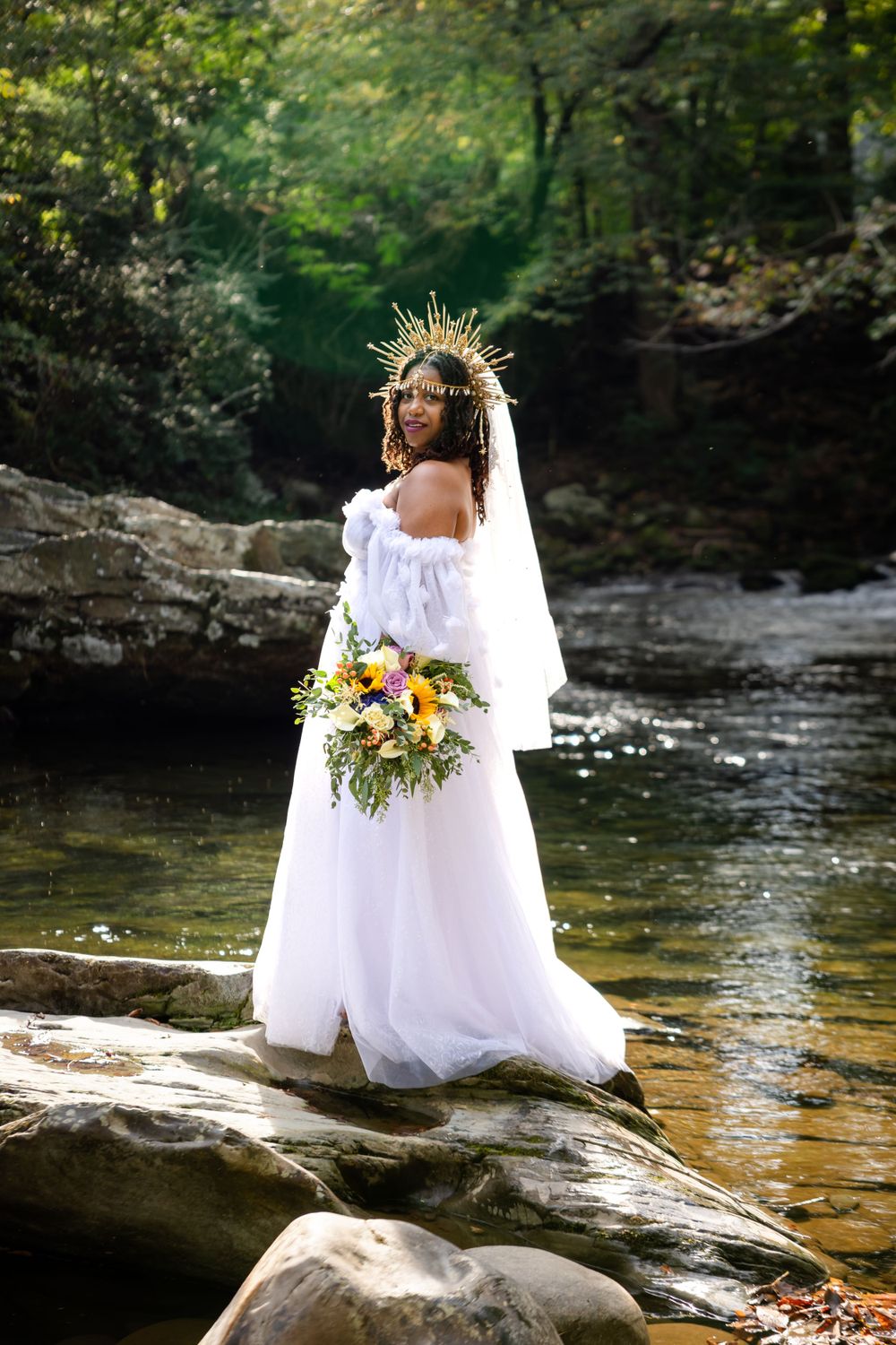 Bride in white dress and crown poses by stream in forest setting.