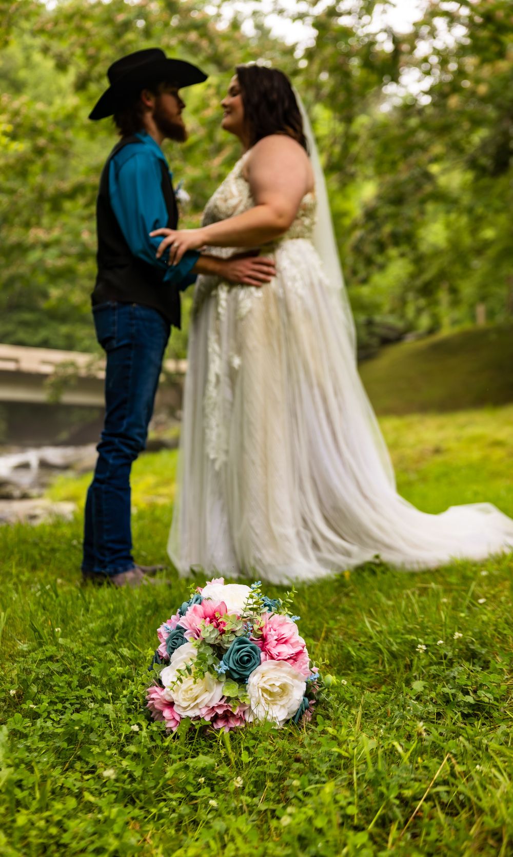 A bride in a white gown and groom in cowboy hat stand on green grass, with a pink and teal bouquet in the foreground.