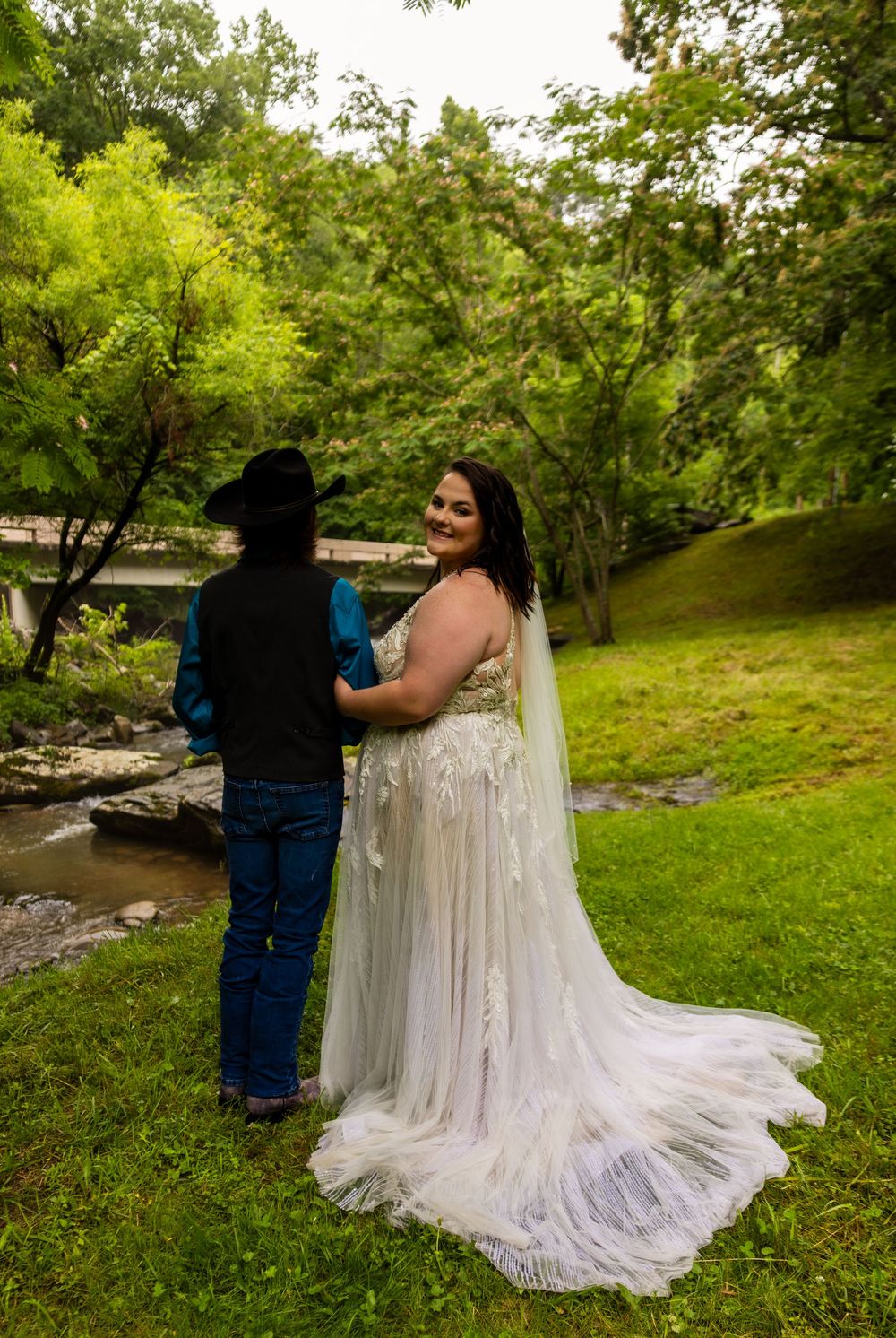 Bride in white lace gown with veil stands with groom in cowboy hat and jeans by a creek in lush greenery.
