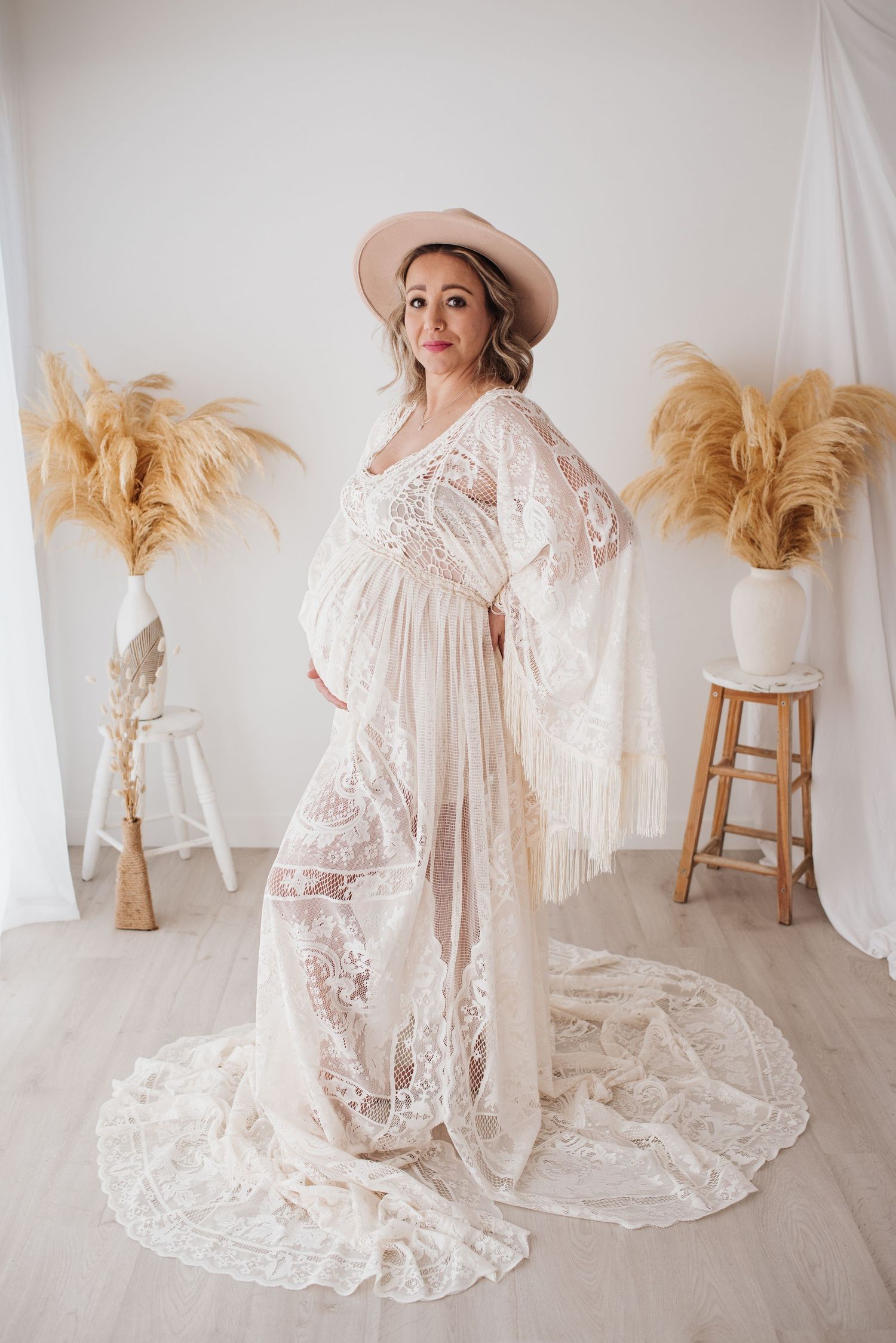 Maternity photo of woman in flowing lace gown and hat posing with pampas grass decor.