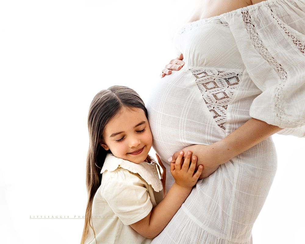Young girl smiling and hugging pregnant mother's belly in a bright white maternity photo session.