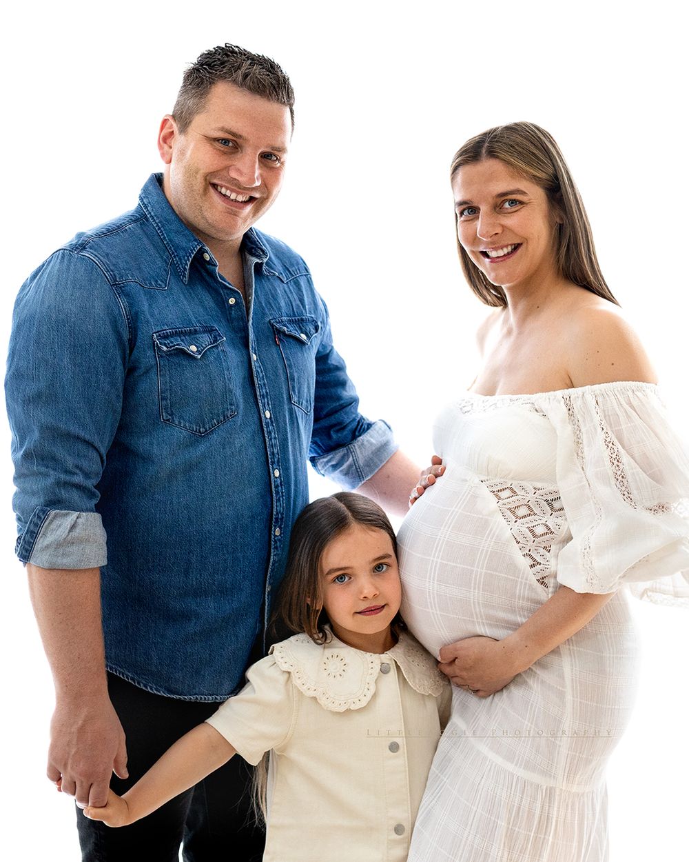 Smiling family maternity photo with man in denim shirt, pregnant woman in white dress, and young girl on white background.