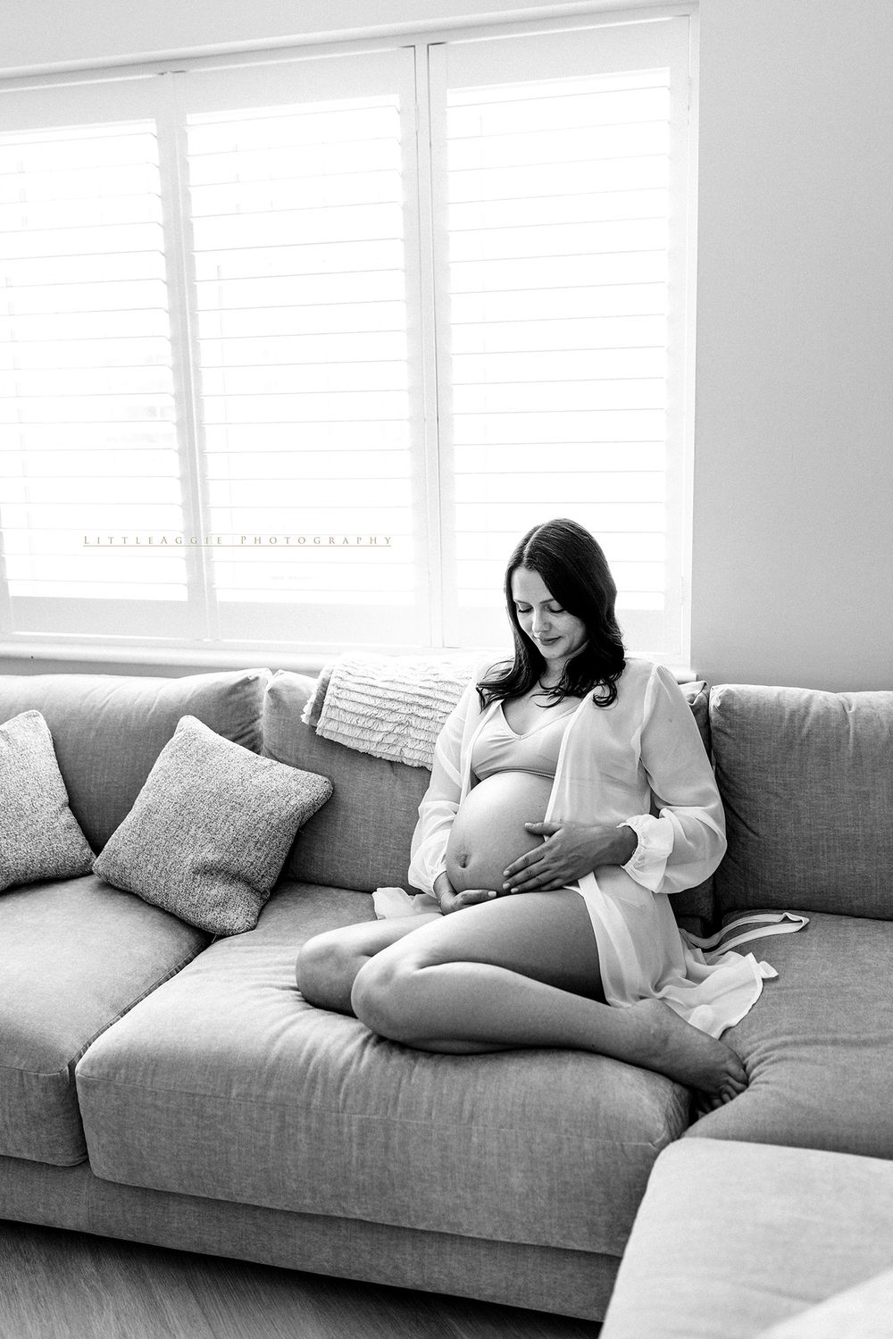 Black and white photo of a pregnant woman sitting cross-legged on a sofa, cradling her baby bump near a bright window.