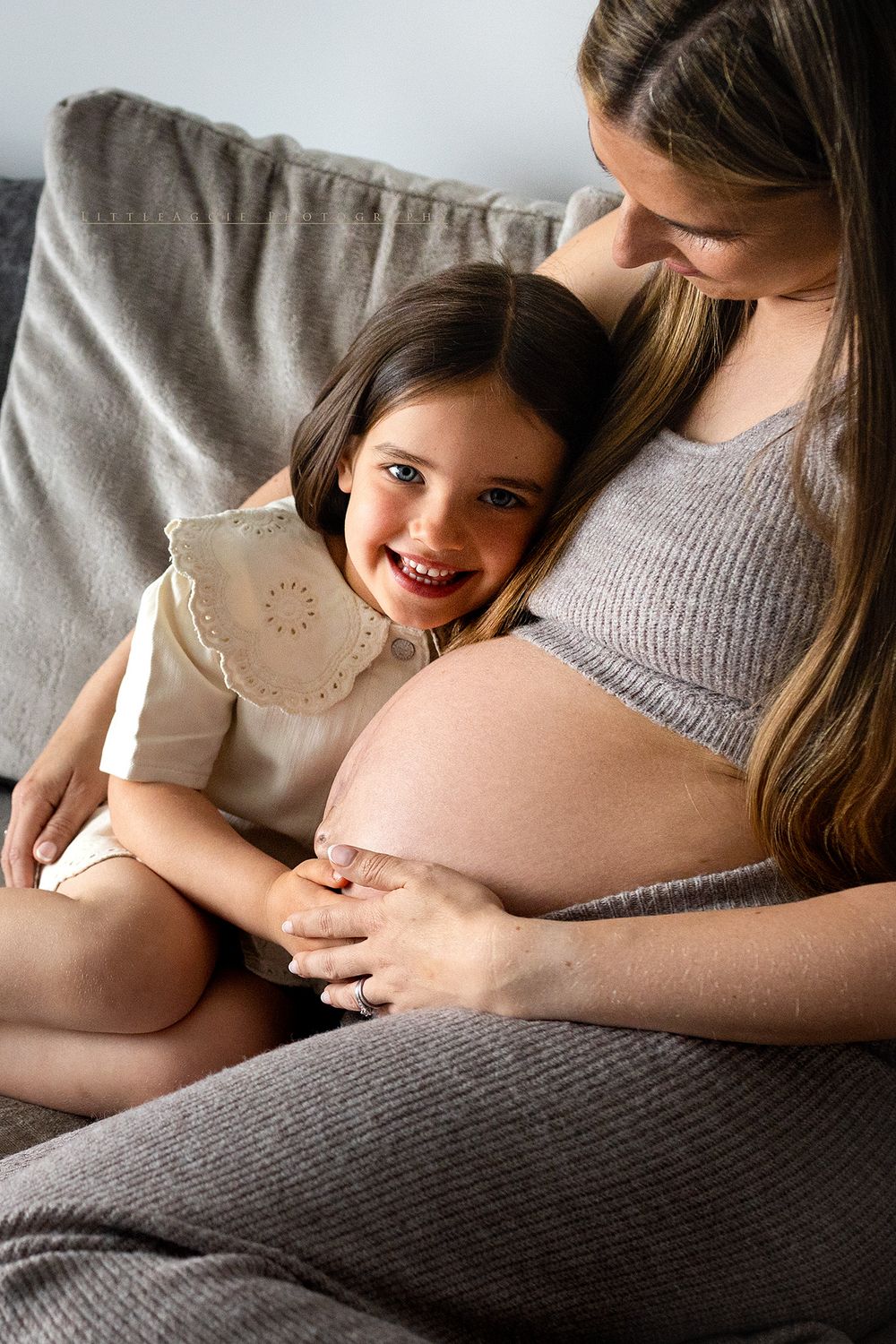 A smiling young girl rests against her pregnant mother's belly while sitting together on a gray sofa at home.