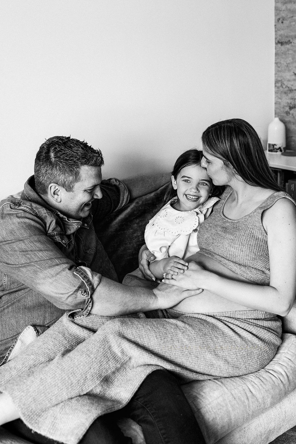 Black and white photo of a happy family with pregnant mother, father, and young child sitting together on a couch.