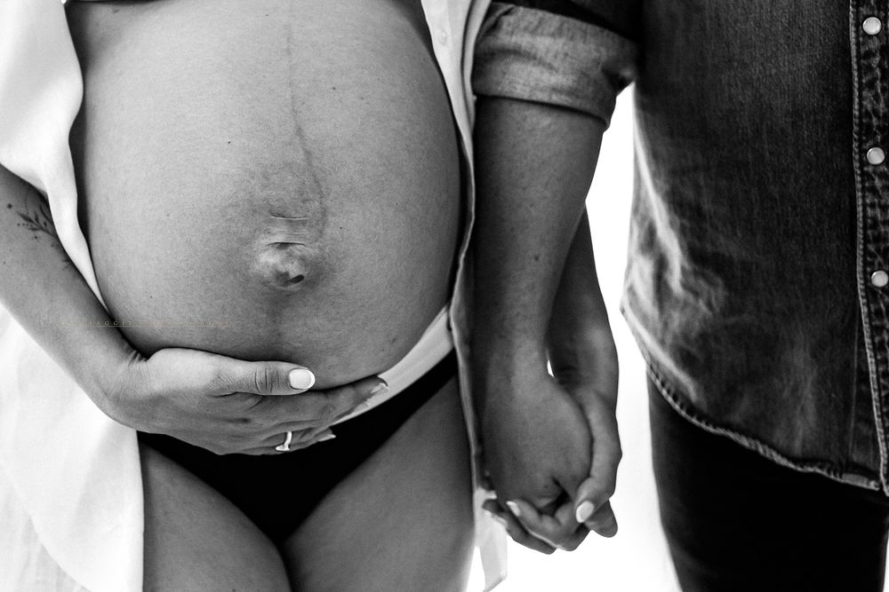 Black and white maternity photo of pregnant woman holding belly while partner holds her hand.