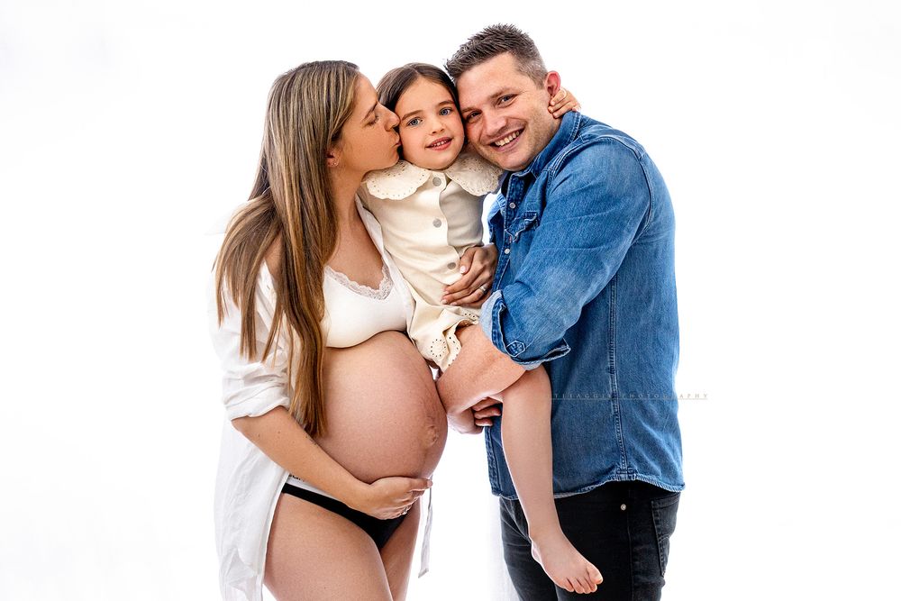Pregnant woman and man holding toddler girl, smiling family maternity photo shoot on white background.