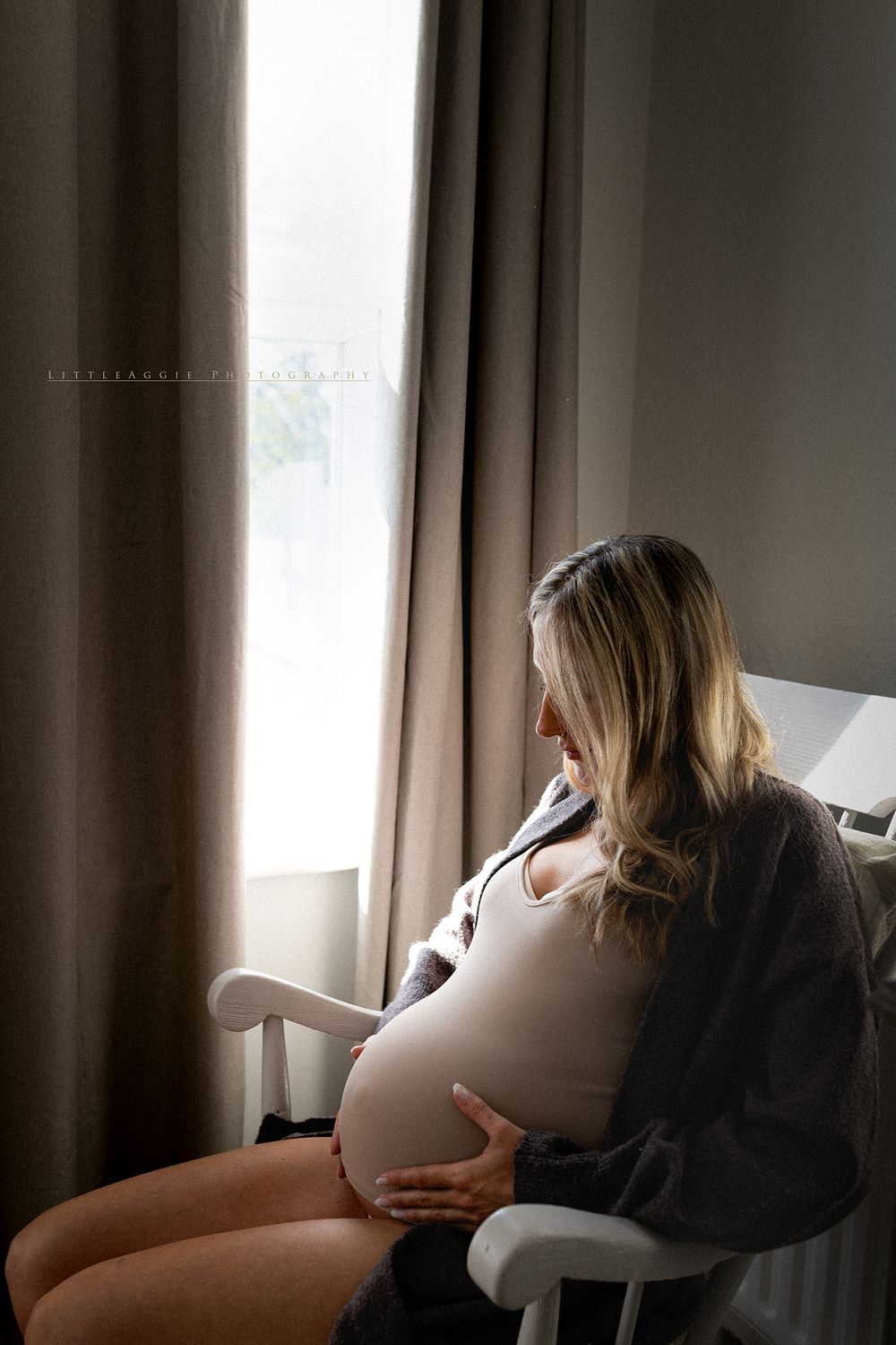Pregnant woman sitting by window cradling baby bump, wearing white bodysuit and gray robe in soft natural light.